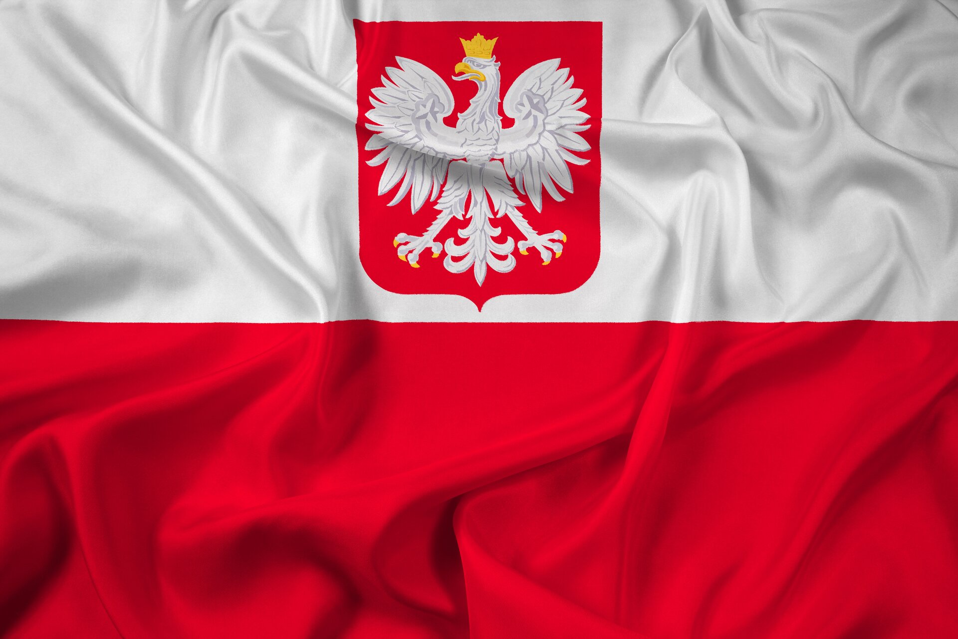 Ilustracja przedstawia flagę z herbem Polski. Cały kadr zdjęcia wypełnia biało‑czerwona, pofałdowana flaga. Na białej części umieszczony jest herb z orłem w koronie.