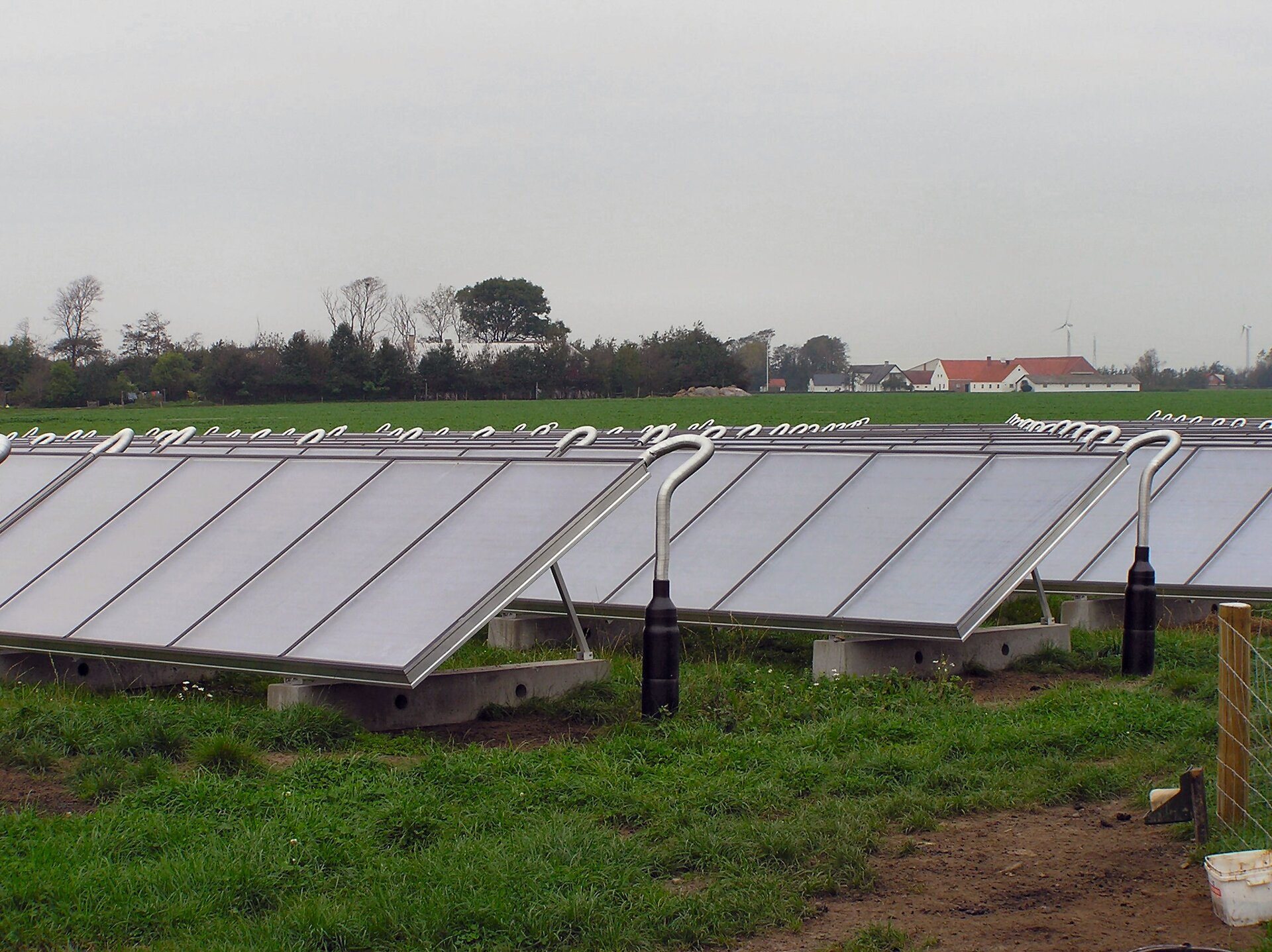 Na ilustracji widać trzy rzędy paneli słonecznych ustawionych na trawiastym polu. W oddali znajdują się po prawej stronie zabudowania gospodarcze a po lewej rosną drzewa i krzewy. W dalekim planie widać zarysy wiatraków