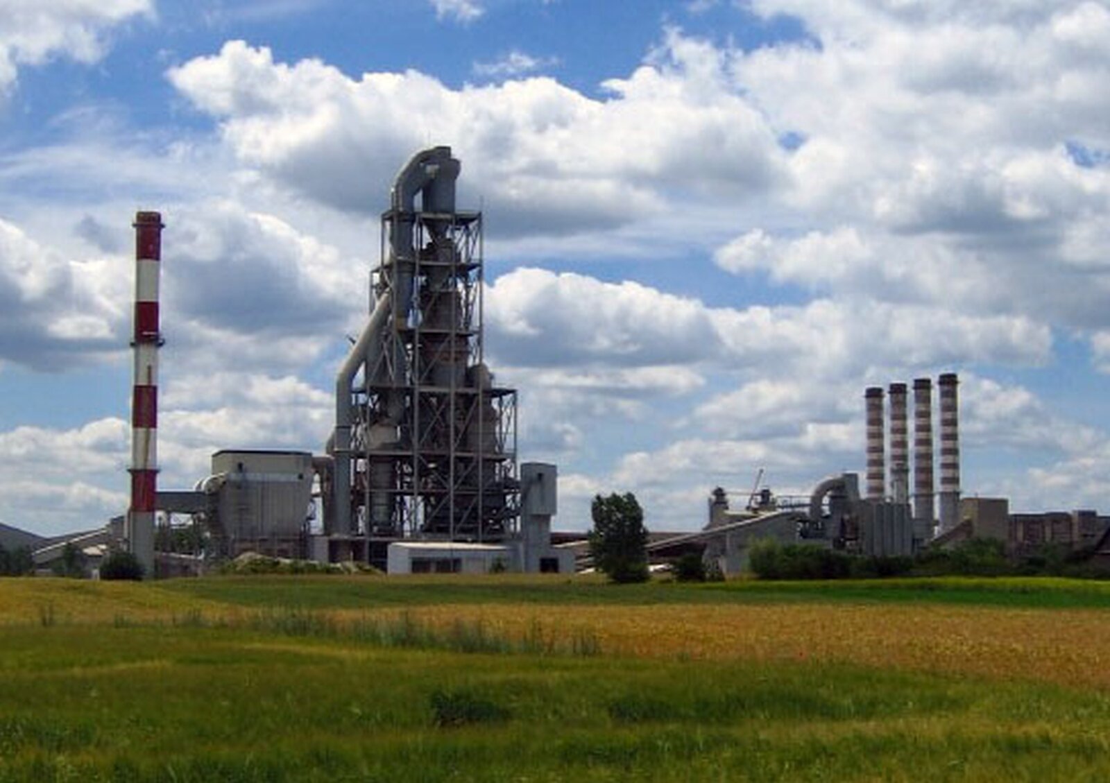 Na zdjęciu budynki przemysłowe, wokół których wybudowane są wysokie kominy. Przed przemysłową infrastrukturą pola uprawne porośnięte zieloną i pomarańczową niską roślinnością.