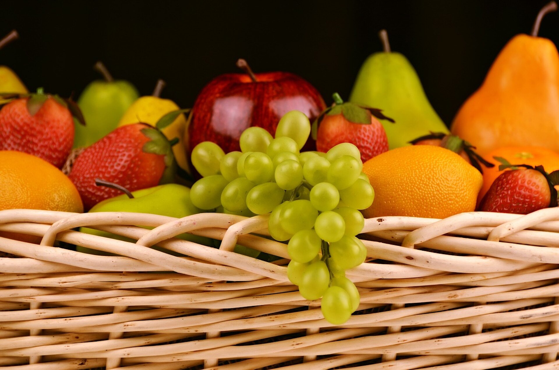 Zdjęcie przedstawia wiklinowy kosz z owocami: truskawki, kiść winogron, gruszki, pomarańcze i jabłka.