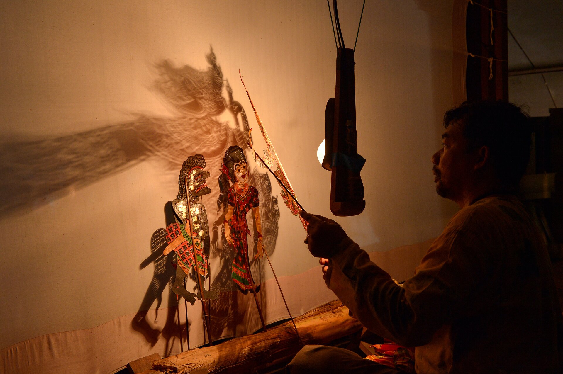 Ilustracja przedstawia prace lalkarza w teatrze cieni. Na fotografii widoczny jest mężczyzna trzymający szablony, których cienie odbijają się na ścianie, nad ustawionymi na jej tle lalkami. Scena dzieje się na tle rozpiętego, białego płótna, przed którym zawieszona jest lampa.