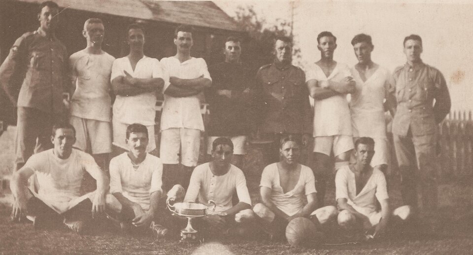 Zdjęcie przedstawia drużynę, która pozuje do zdjęcia. Składa się z dwóch rzędów młodych mężczyzn. Pierwszy rząd siedzi na trawie, a drugi stoi. Większość jest ubrana w jasne stroje sportowe, a kilku w mundury. Przed drużyną stoi puchar.