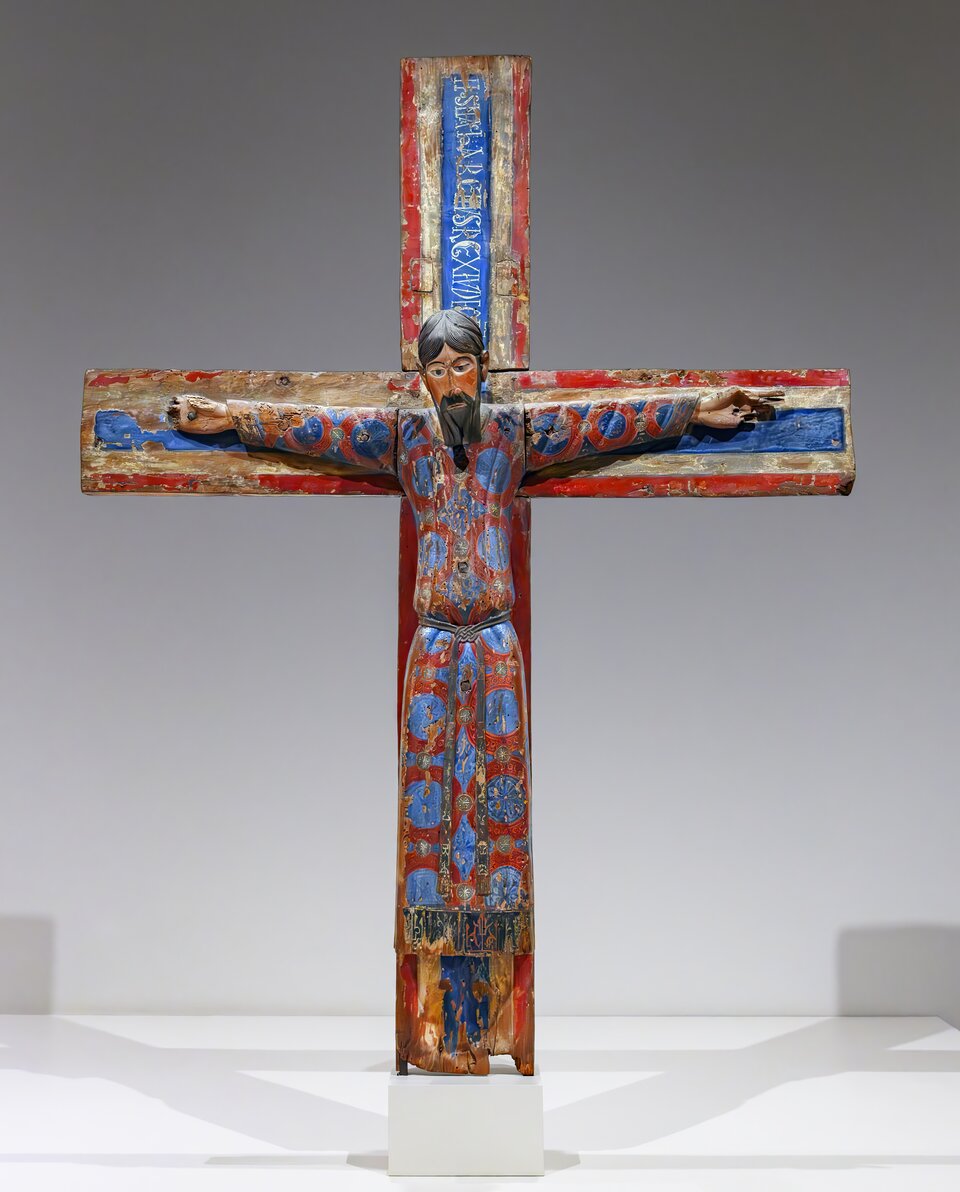 Romański, polichromowany krucyfiks: Chrystus ukazany frontalnie na drewnianym krzyżu, z rozłożonymi ramionami, ubrany w długą, bogato zdobioną tunikę w barwach czerwieni i błękitu; rzeźba eksponowana na jasnym postumencie w przestrzeni muzealnej.