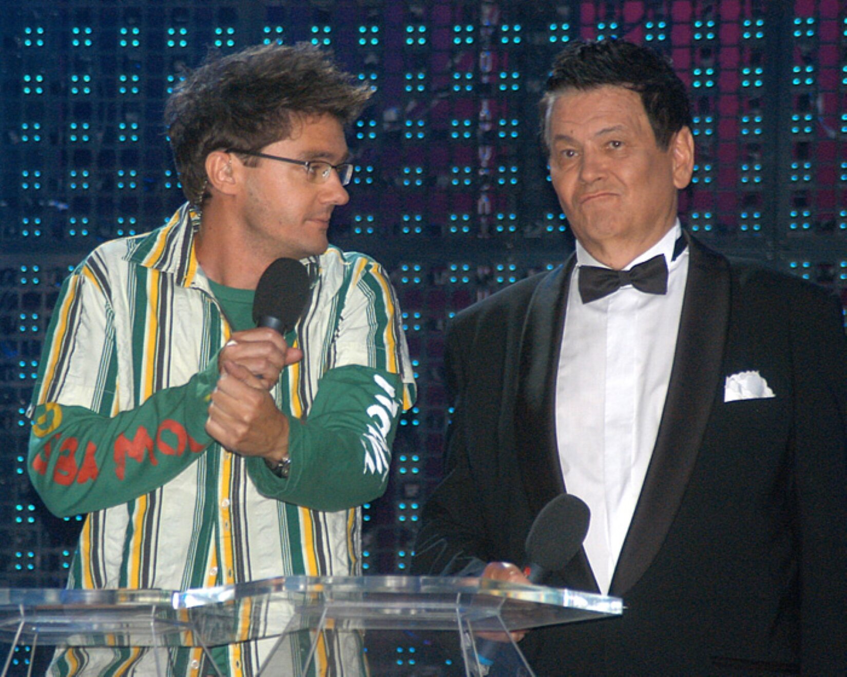 Na zdjęciu stoją dwaj mężczyźni. Mężczyźni znajdują się na granatowym tle z delikatnymi niebieskimi kropeczkami. Przed nimi stoi oszklony stolik. Mężczyzna z lewej strony zdjęcia trzyma obiema dłońmi, blisko twarzy, czarny mikrofon. Ma obróconą twarz w lewą stronę, a na jego nosie znajdują się korekcyjne okulary. Ma czarne włosy, a na sobie ma założoną zieloną koszulę z biało‑żółtymi pionowymi paskami. Na jego lewym przedramieniu, znajduje się czerwony napis, a na prawej ręce biały. Po prawej stronie znajduje się mężczyzna, który jest ustawiony przodem do zdjęcia. W prawej dłoni na wysokości brzucha ma uniesiony czarny mikrofon. Mężczyzna ma skwaszoną minę. Ma czarne włosy, a na sobie ma założoną czarną marynarkę z białą chusteczką włożoną do lewej kieszonki przy piersi. Pod marynarką znajduje się biała koszula oraz czarna mucha. 