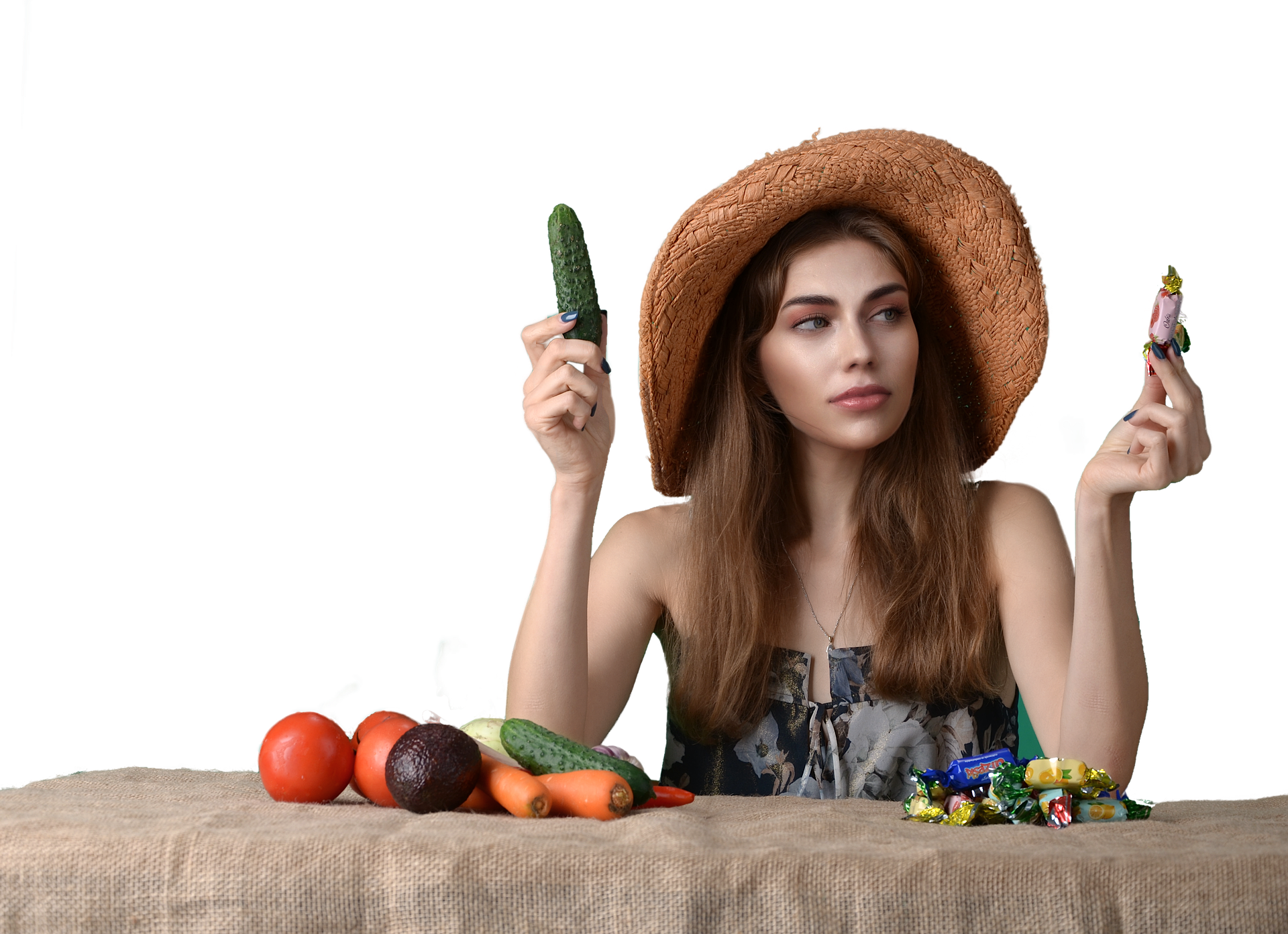 Zdjęcie przedstawia młodą kobietę w kapeluszu na głowie. Przed kobietą leży stos warzyw i cukierków. Kobieta trzyma w jednej dłoni ogórek, a w drugiej cukierek. 