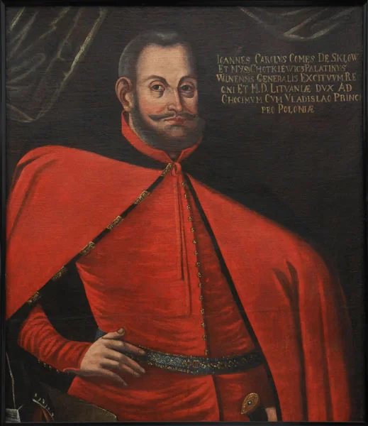 Obraz przedstawia portret mężczyzny w średnim wieku. Mężczyzna ma owalną twarz, ciemne włosy i bujną brodę i wąsy. Na jego twarzy widać rumieńce. Mężczyzna jest ubrany jest w czerwoną pelerynę zarzuconą na kontusz prawą rękę ma opartą na pasie, lewą trzyma na szabli.