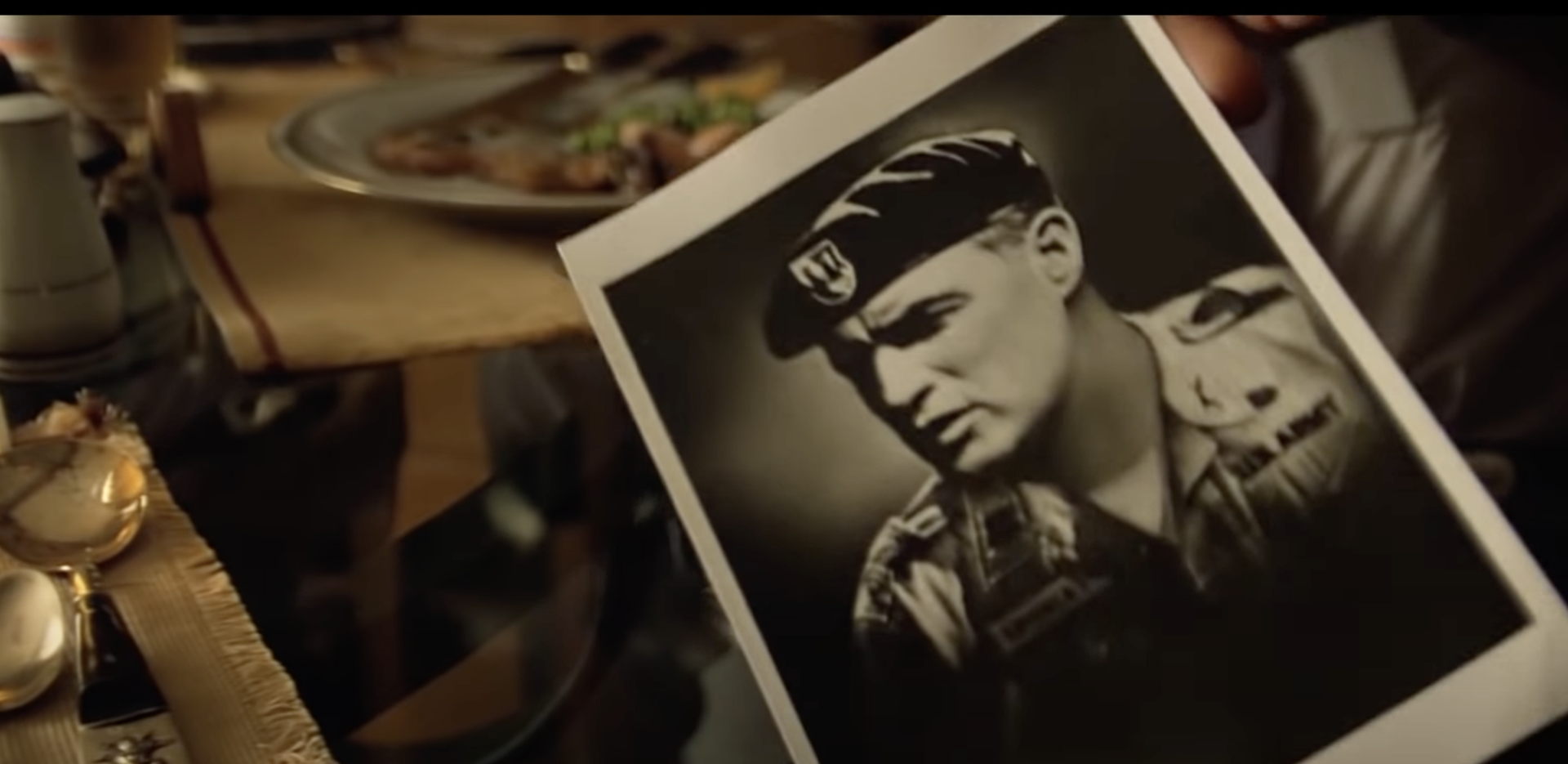 Zdjęcie przedstawia fotografię młodego żołnierza w mundurze i z beretem na głowie. Za fotografią znajduje się stół, na którym są sztućce, szklanki i talerz z jedzeniem.