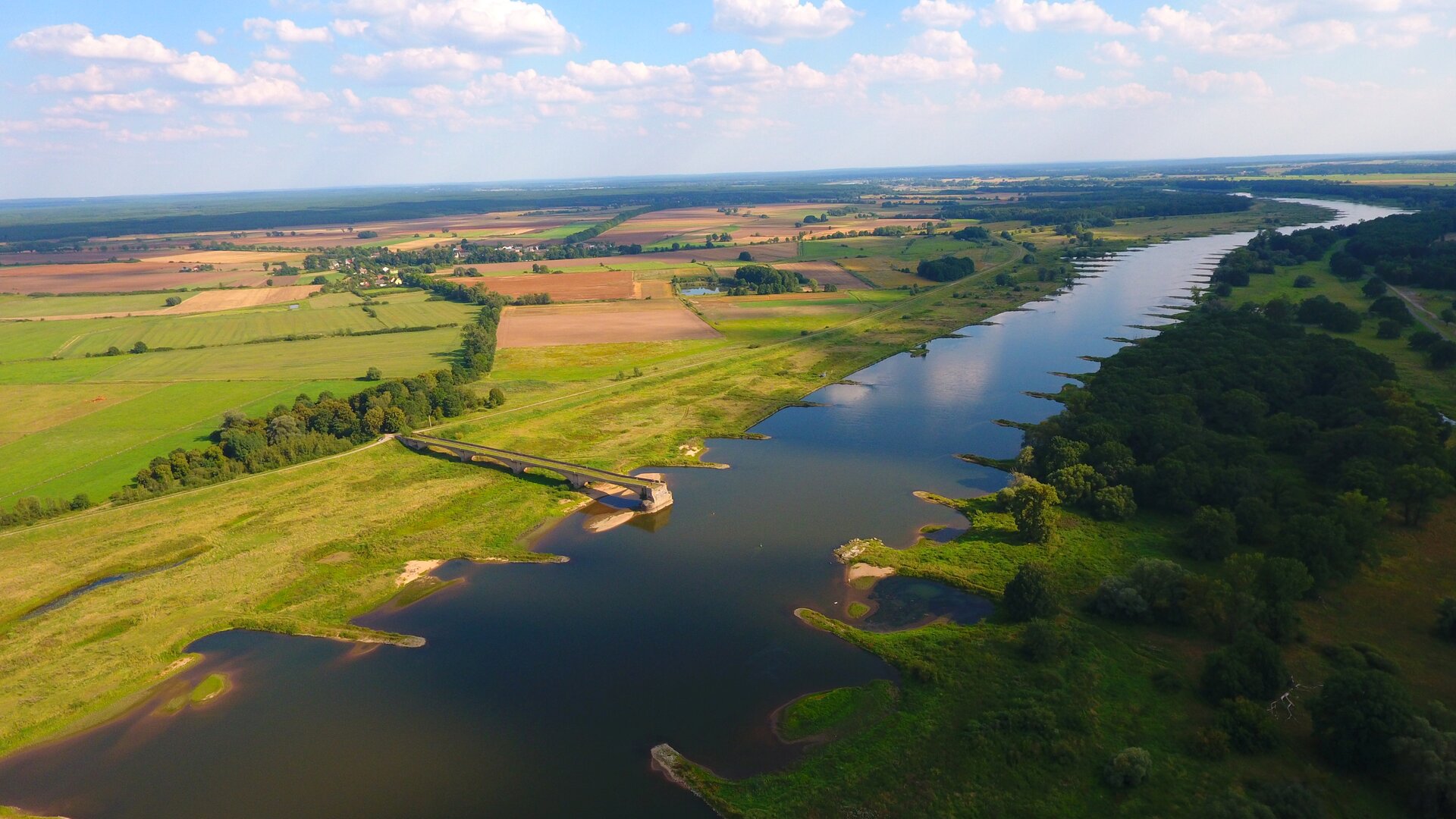Lotnicza fotografia doliny rzeki. Dookoła płaski, rozległy teren pokryty lasami i polami uprawnymi.