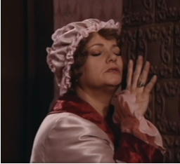 Zdjęcie przedstawia kobietę z zamkniętymi oczami, która dotyka twarzą pieca kaflowego. Ma na głowie różowy czepek, ubrana jest w różową podomkę z czerwonym kołnierzem.