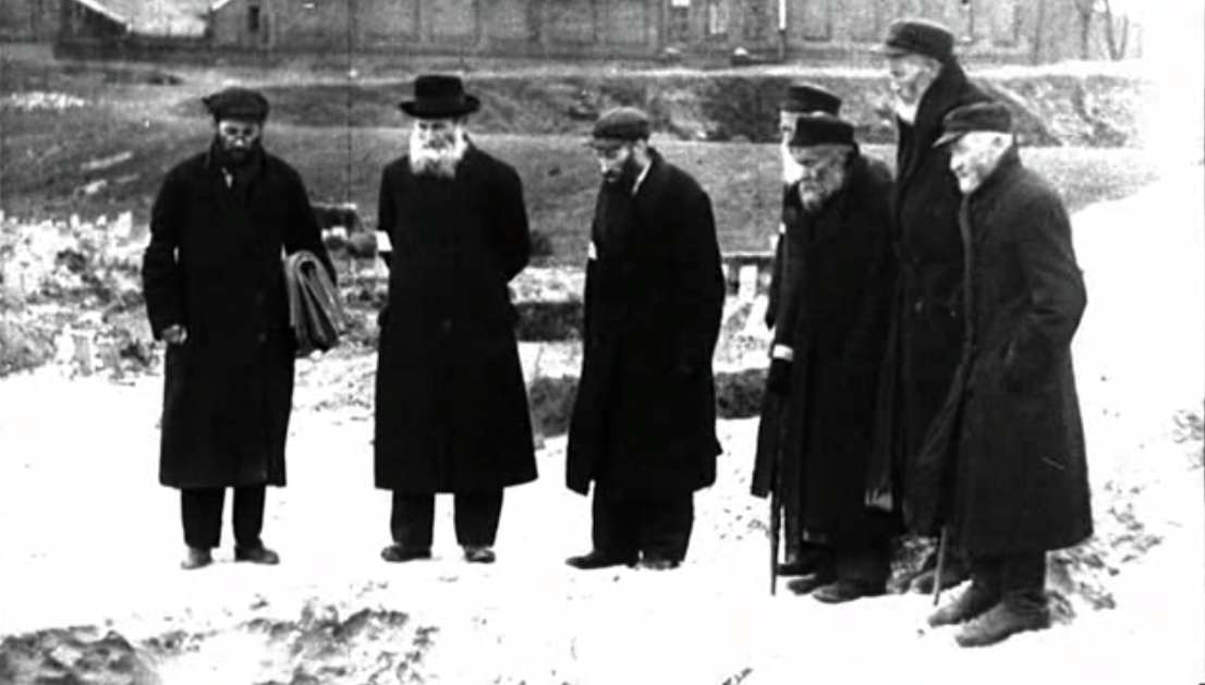 Zdjęcie przedstawia sześcioro mężczyzn w starszym wieku. Ubrani są w długie, ciemne płaszcze oraz kapelusze. Mężczyźni mają długie brody. Wpatrują się z pochylonymi głowami w doły wykopane w ziemi.