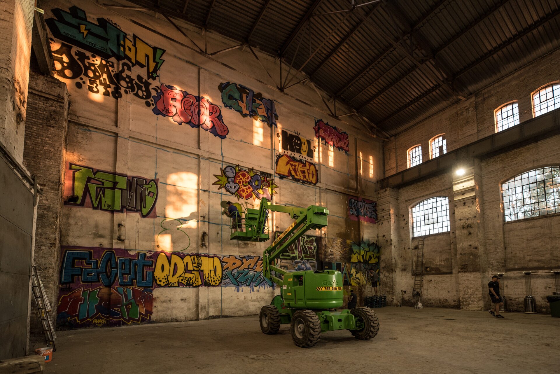 Na zdjęciu znajduje się wysokie pomieszczenie o jasnych, surowych ścianach i podłodze. Na ścianie na wprost jest wiele kolorowych graffiti [czytaj: grafiti], a na ścianie po prawej stronie jest 6 okien ze szprosami. Na środku pomieszczenia stoi zielony podnośnik koszowy. Na fotografii są dwie osoby malujące graffiti, jedna w koszu podnośnika, druga stojąca na stosie z palet.