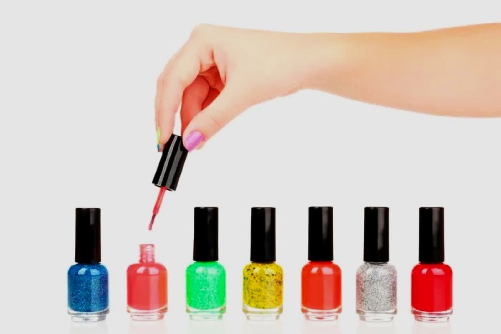 Zdjęcie przedstawia siedem słoiczków z lakierami do paznokci, stojących w rzędzie. Nad drugim z kolei, otwartym unosi się ręka kobiety trzymająca nakrętkę z pędzelkiem.
