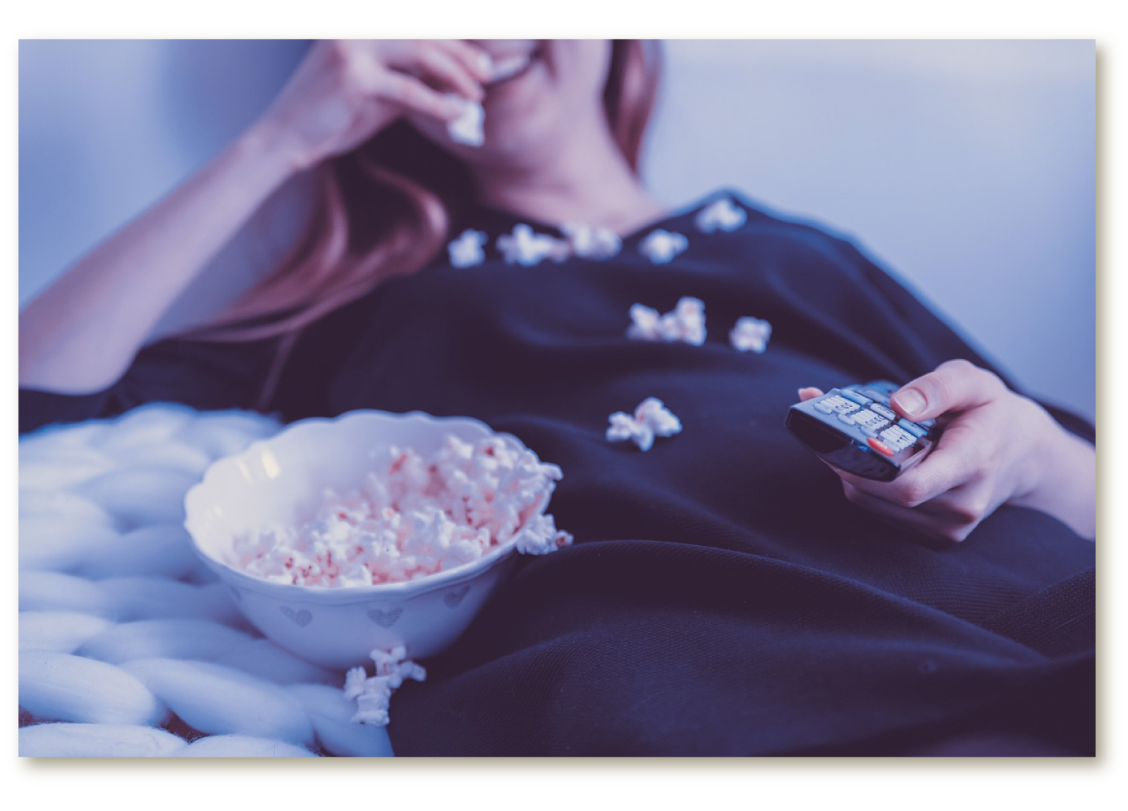 Zdjęcie przedstawia leżącą dziewczynę. Obok niej miska z popcornem. Dziewczyna jedną ręką trzyma pilota od telewizora, a drugą je popcorn. 