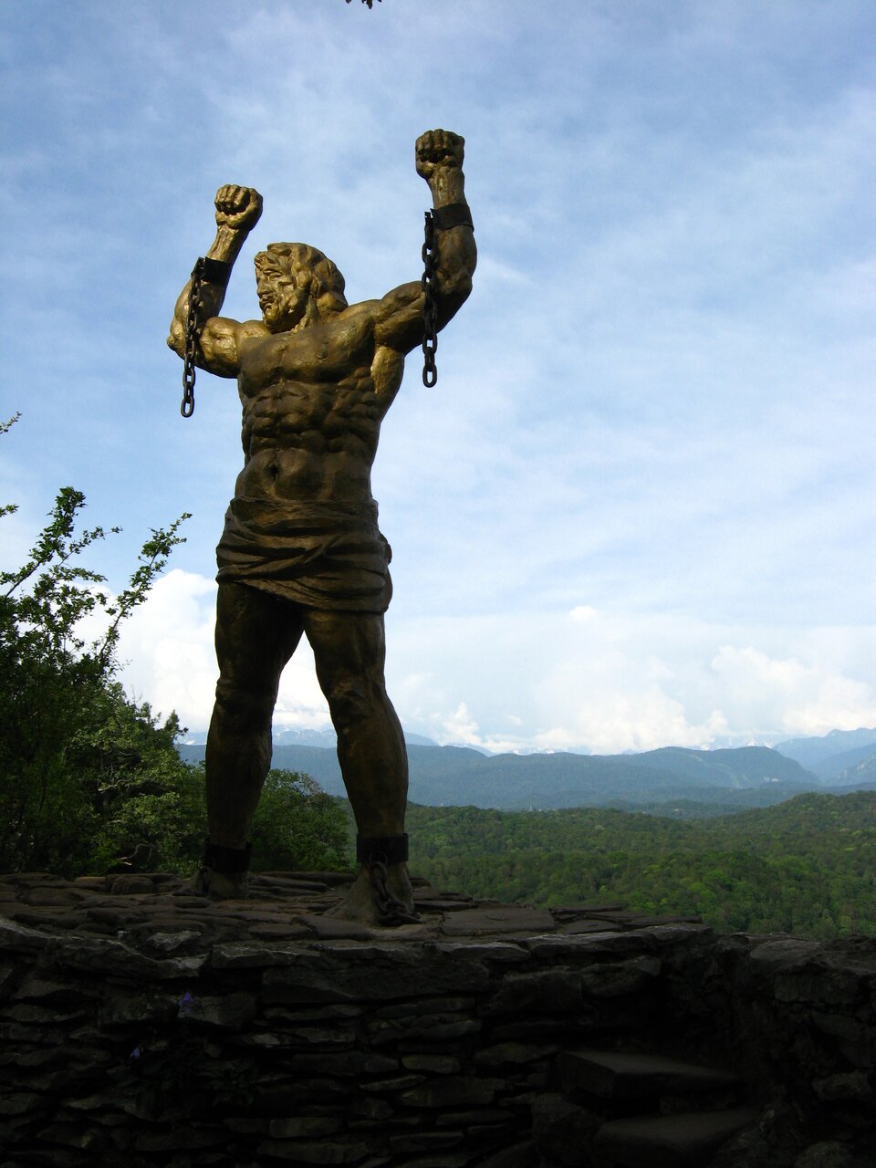 Zdjęcie przedstawia rzeźbę mężczyzny z uniesionymi dłońmi. Mężczyzna ubrany jest jedynie w szatę przewiązaną na jego biodrach. Ciało mężczyzny jest umięśnione. Mężczyzna wznosi ręce z zaciśniętymi pięściami ku górze. Na nadgarstkach mężczyzny widoczne są kajdany z fragmentem łańcucha, który został zerwany. Rzeźba wykonana została z ciemnego, brązowego materiału. Stoi ona na fragmencie skalnego wzniesienia. Obok widoczny jest fragment zielonych krzewów. W tle za rzeźbą widoczny jest las i czubki drzew. Niebo jest błękitne i bezchmurne. Zdjęcie zostało wykonane w świetle dziennym.