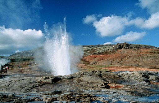 Ilustracja przedstawia Geysir - jeden z najbardziej znanych islandzkich gejzerów. Woda potrafi wypryskiwać z niego nawet na kilkadziesiąt metrów.  Wokół rozciąga się wzgórze, widać błękitne niebo z jasnymi chmurami.