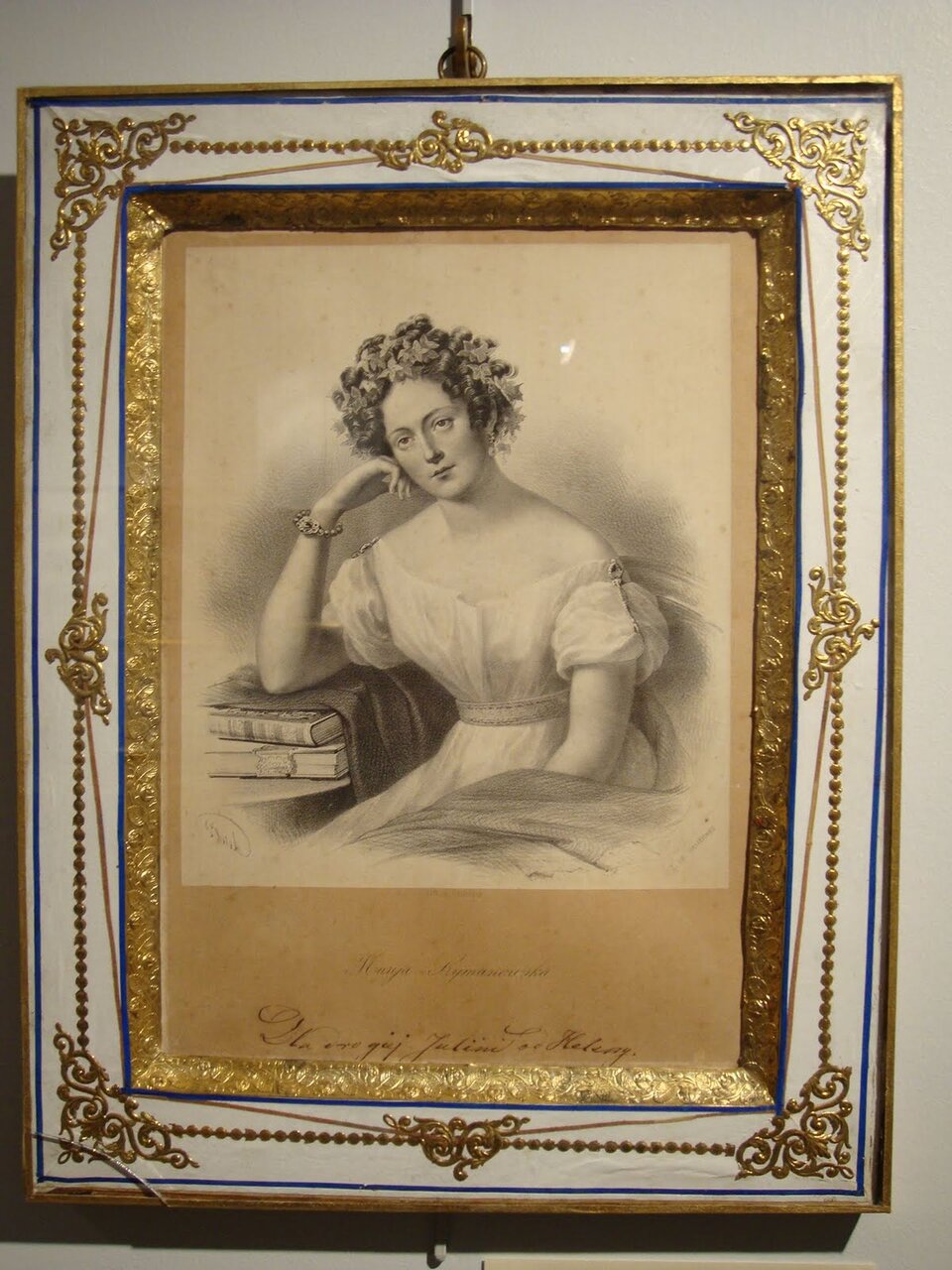 Oprawiony w ozdobną ramę portret kobiety w stylu XIX‑wiecznym. Grafika w odcieniach sepii przedstawia młodą kobietę siedzącą przy stole, opartą głową na dłoni w zamyślonej pozie. Ma na sobie lekką, jasną suknię z krótkimi rękawami i ozdobną opaskę z kwiatów we włosach. Na ręce widoczna jest bransoletka. Obok niej leżą książki, co sugeruje intelektualny lub literacki charakter przedstawienia. Całość otoczona jest bogato zdobioną ramą z elementami złocenia i dekoracyjnymi ornamentami w narożnikach. Pod ilustracją znajduje się podpis, częściowo czytelny.