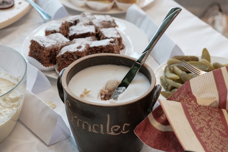 Zdjęcie przedstawia stół przystrojony białym obrusem. W centralnym jego punkcie znajduje się duży, brązowy pojemnik wypełniony białą substancją podpisany „smalec”. W tle znajdują się ogórki kiszone oraz ciasto posypane cukrem pudrem.