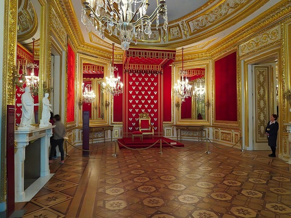 Zdjęcie przedstawia pomieszczenie audiencyjne króla z wyeksponowanym tronem z podnóżkiem, kory znajduje się przy środkowej ścianie. Na ścianie za tronem wisi zaplecek tronu, zdobiony haftowanymi srebrem na tle z czerwonego aksamitu. Wokół tronu są wielkie lustra w złoconych ramach. Przy lewej ścianie stoi marmurowy kominek,  zdobiony antycznymi rzeźbami. W prawej ścianie umieszczone są drzwi. Są otwarte i wchodzi przez nie człowiek. 