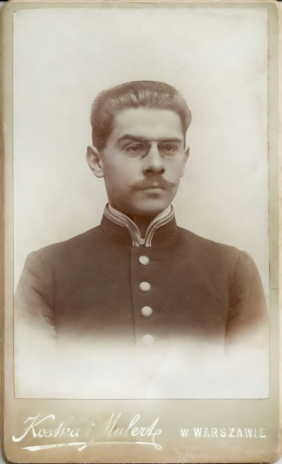 Czarno‑biała fotografia. Mężczyzna  o zaczesanych do tyłu, ciemnych włosach, z wąsem, w binoklach. Ubrany w ciemną marynarkę ze stujką. 