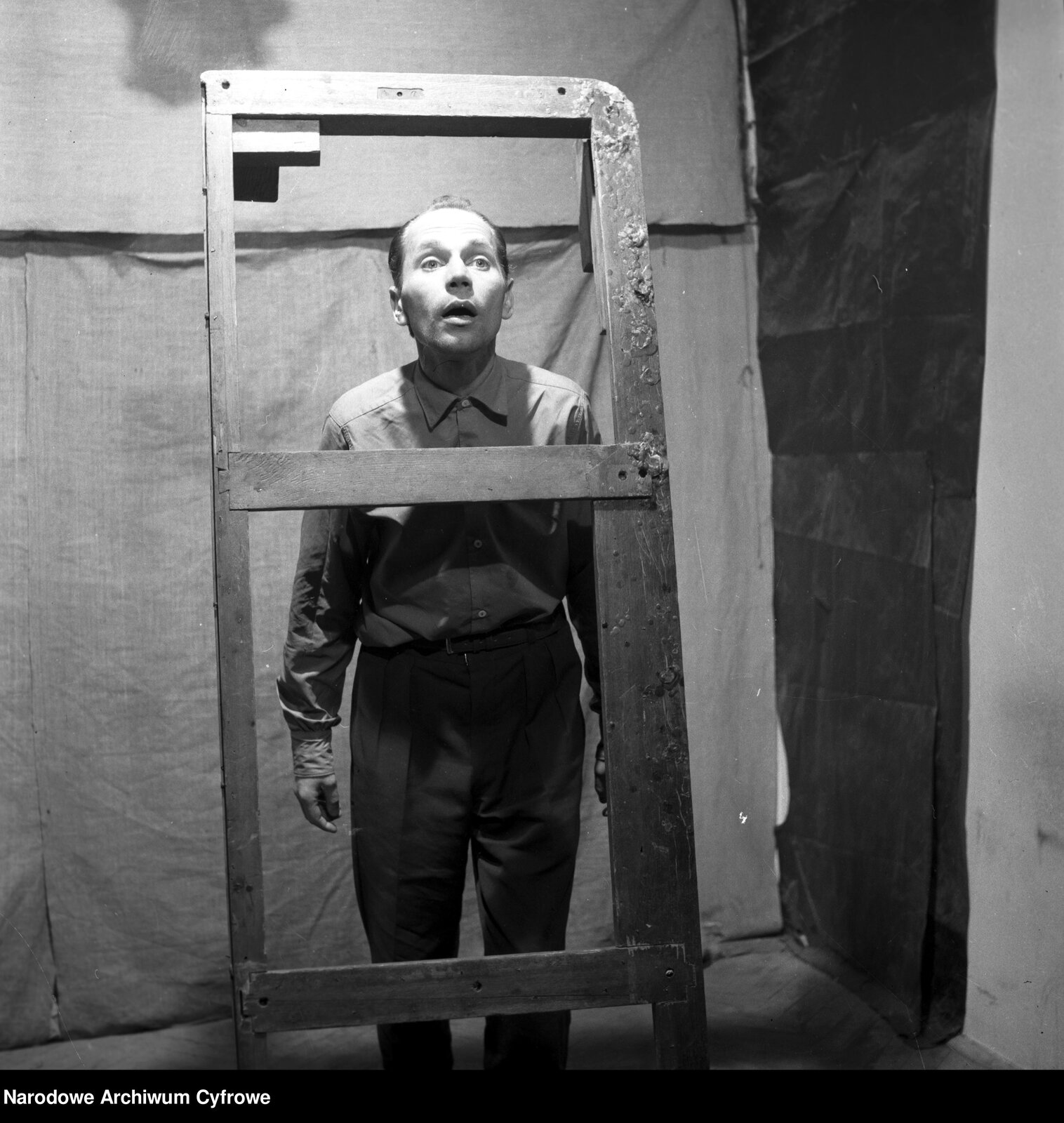 Obraz przedstawia dojrzałego mężczyznę stojącego ma tle płachty materiału. Mężczyzna stoi z opuszczonymi rękami, ma lekko uchylone usta. Ubrany jest w koszulę i spodnie. Stoi za stelażem podstawy szafy.