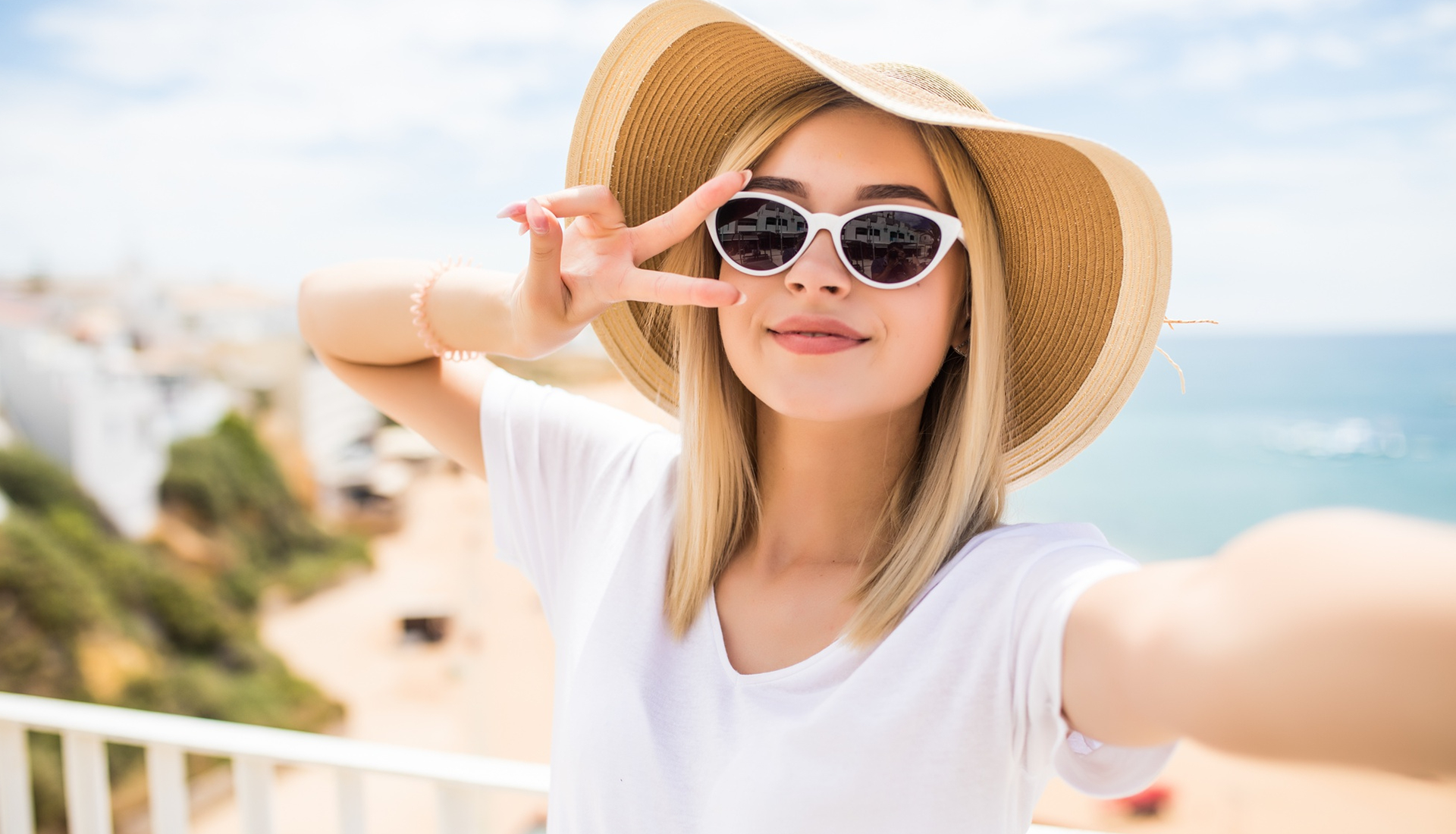 Zdjęcie przedstawia młodą dziewczynę od pasa w górę. Ubrana jest w białą koszulkę z krótkim rękawem. Ma długie, rozpuszczone blond włosy i uśmiechniętą twarz. Na głowie ma duży słomkowy kapelusz, a na twarzy okulary przeciwsłoneczne w białych oprawkach. Prawą rękę ma wyprostowaną i uniesioną lekko do góry. Lewą rękę ma zgiętą i podniesioną do góry na wysokość twarzy. Palce wskazujący i środkowy ułożone są w literę V. W tle widoczna jest rozmyta plaża, woda i wybrzeże z zabudowaniami. 
