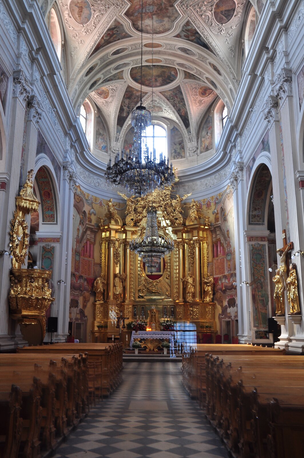 Wnętrze Kościoła pw św. Franciszka Ksawerego w Piotrkowie Trybunalskim. Na fotografii znajduje się widok na ołtarz, nawę główną oraz sklepienie. Ołtarz zajmuje całą szerokość nawy głównej, jest bogato złocony i rzeźbiony. Na ścianie bocznej kościoła widoczna jest złota ambona po lewej stronie, a po prawej – krucyfiks pomiędzy dwoma złotymi posągami. Sklepienie jest wypełnione malowidłami, zwisają z niego również kryształowe żyrandole. Tuż pod sklepieniem umieszczone są otwory okienne zwieńczone półokrągłym łukiem. W nawie znajdują są także drewniane ławki stojące w dwóch rzędach.