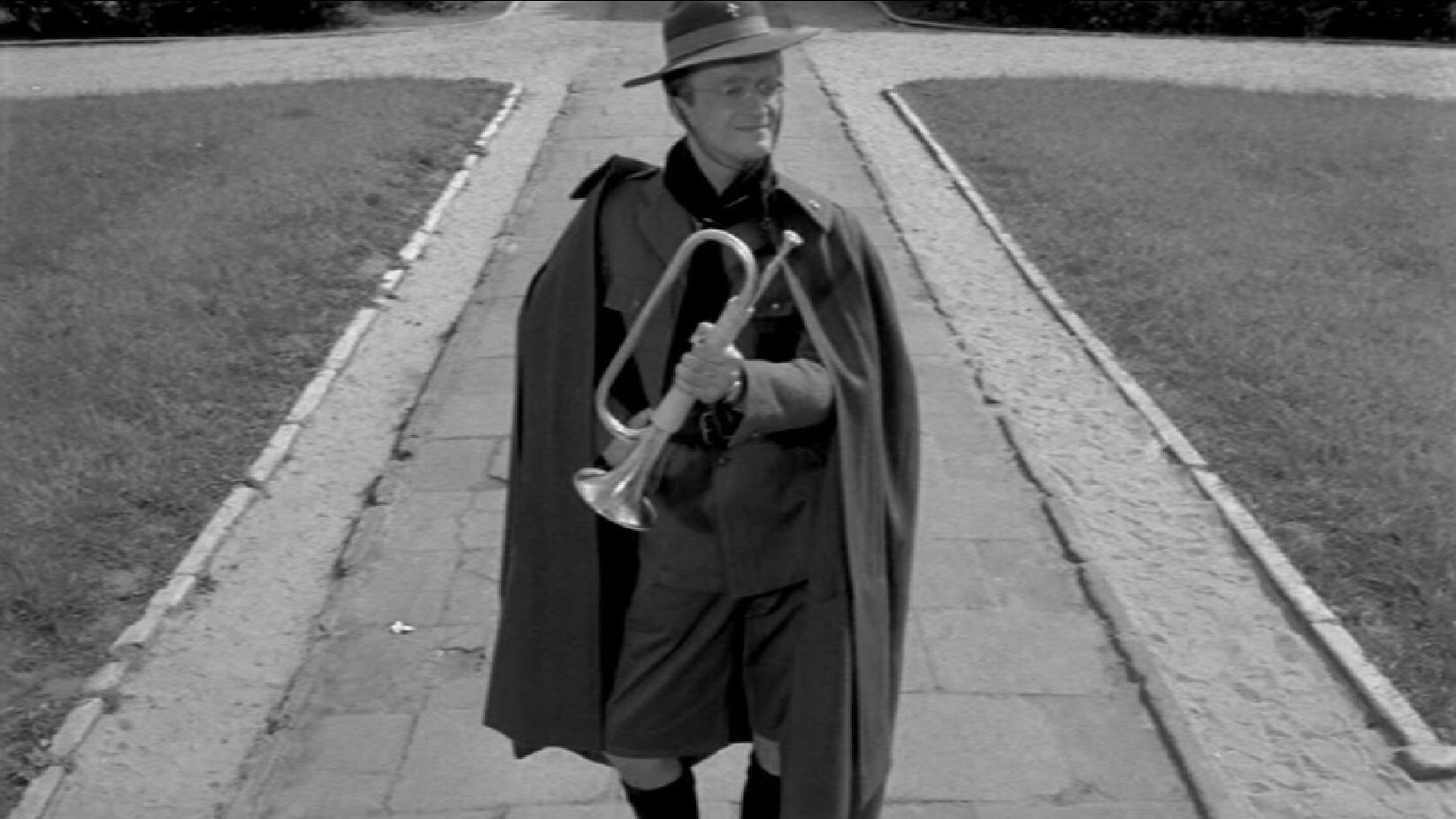 Zdjęcie przedstawia mężczyznę w stroju harcmistrza, z peleryną na ramionach i kapeluszem na głowie. Mężczyzna trzyma w dłoniach trąbkę. 