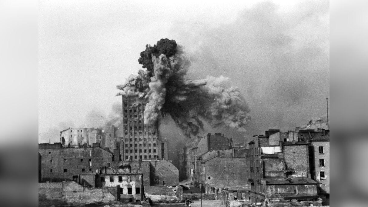 Zdjęcie przedstawia fragment zabudowań miasta. W środku wysoki kilkunastopiętrowy budynek pokazany w momencie trafienia przez pocisk. Górna część budynku wybucha.
