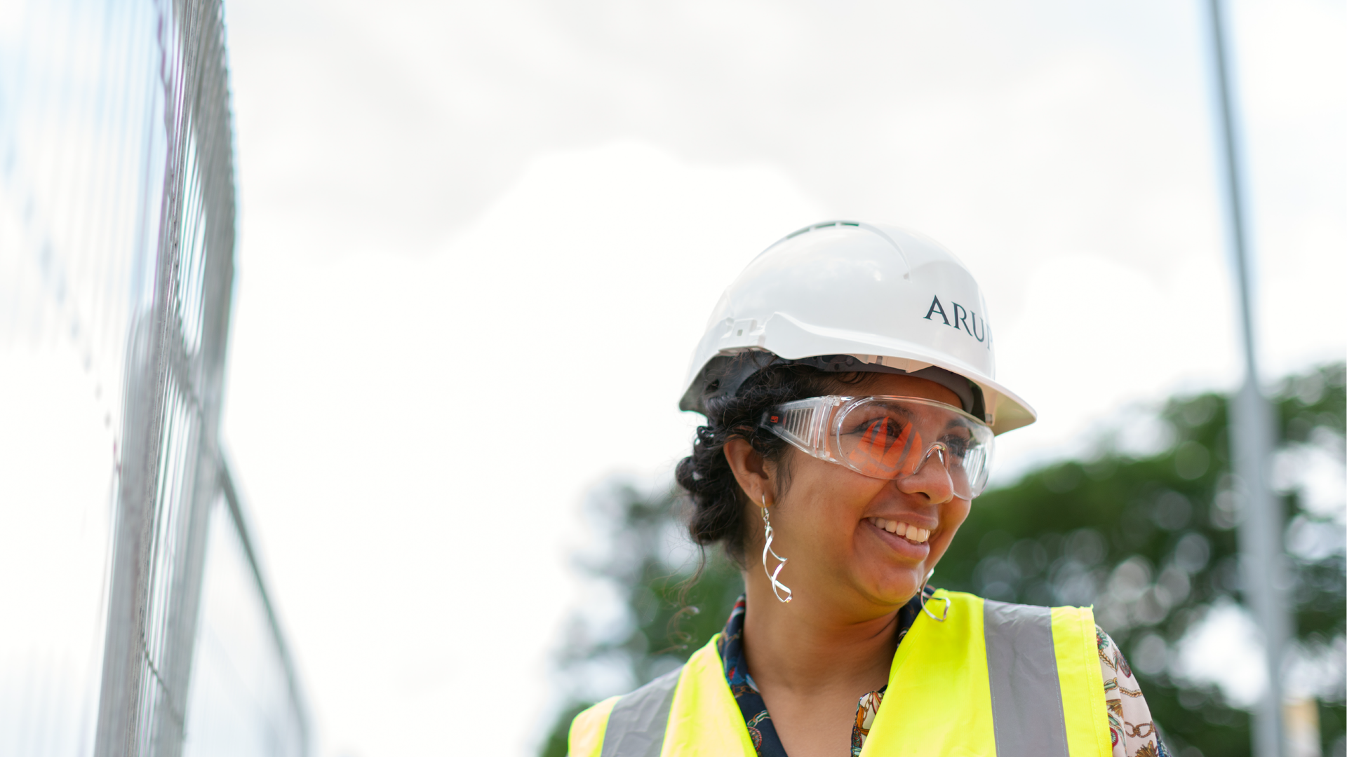 Zdjęcie przedstawia uśmiechniętą, czarnoskórą dziewczynę. Na głowie ma ochronny kask, na oczach ochronne okulary. Na sobie ma również kamizelkę odblaskową.