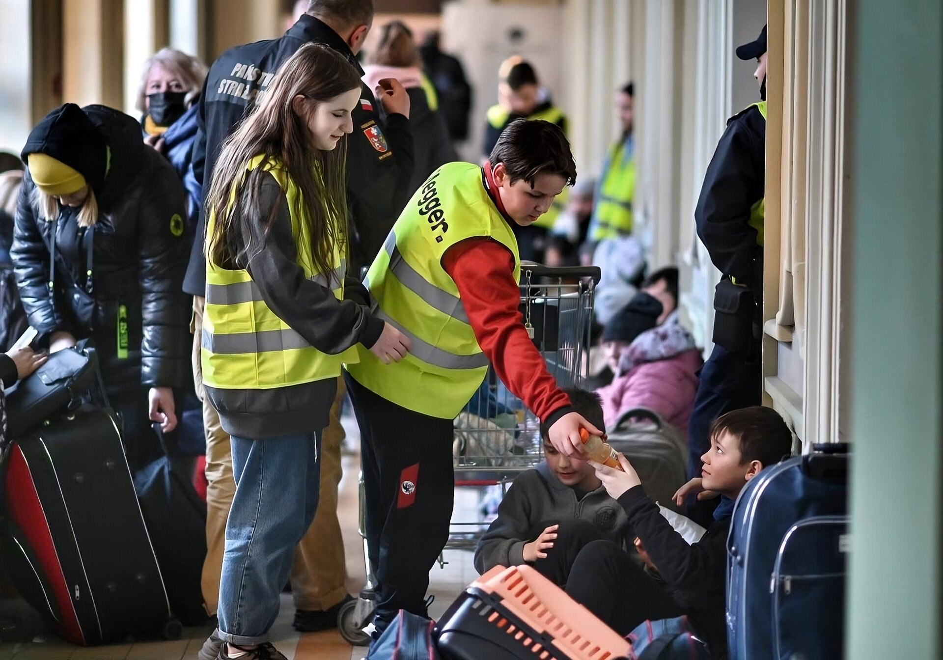 Kliknij, aby powiększyć Zdjęcie przedstawia ciasno siedzących z walizkami, torbami podróżnych. Po lewej stronie kobieta schyla się i podnosi swoje bagaże. Po prawej stronie siedzi dwoje dzieci obstawionych bagażami. Nad nimi stoi dwóch wolontariuszy - dziewczynka i chłopiec. Chłopiec podaje soczek. W oddali widać funkcjonariuszy straży granicznej.
