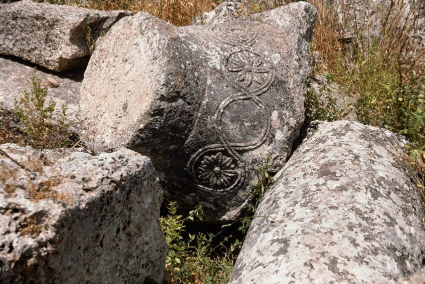 Na zdjęciu górna część kolumny. Leży na trawiastym podłożu, wśród ruin. Wykonana jest z kamienia. Znajdują się na niej spiralne wzory z elementami kwiatowymi. 