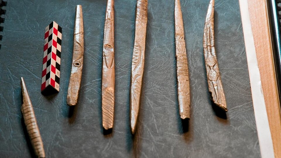 Ilustracja przedstawia sześć wyrobów z kości, ułożonych w pionie na ciemnym tle, jedna obok drugiej. Z jednej strony są zakończone owalnie, z drugiej są szpiczaste. Na ich powierzchni znajdują się nacięcia, tworzące geometryczne wzory. 