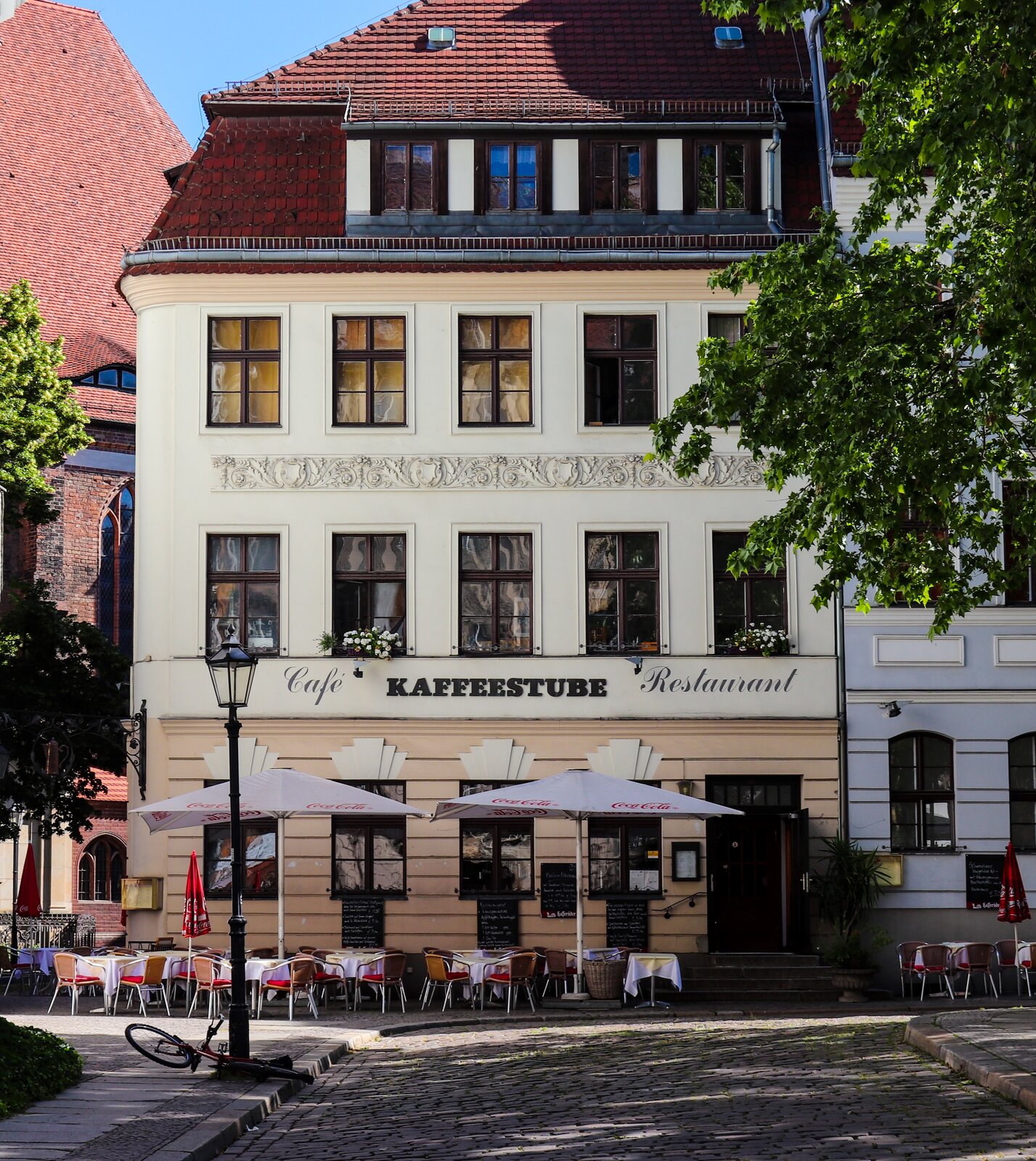 Zdjęcie przedstawia brukowaną ulicę, przy której stoi duża, czterokondygnacyjna kamienica. Na budynku znajduje się napis: Cafe, Restaurant, Kaffestube. Przed budynkiem stoją pod dwoma parasolami stoliki i krzesła.