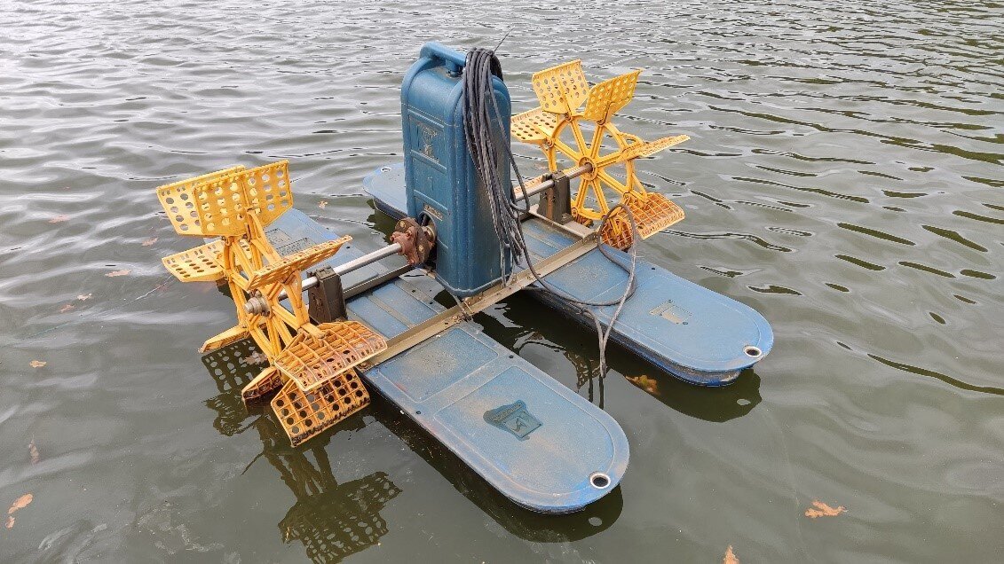 Zdjęcie przedstawia aerator z bliska. Pływa on na dwóch bojkach. Od jego środkowego elementu odchodzą w bok rurki, na końcach których znajdują się łopatki.