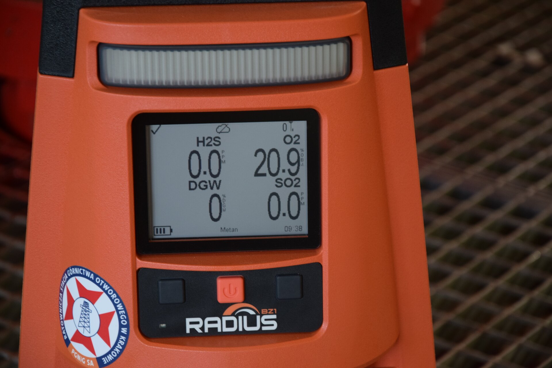 Na zdjęciu widoczne jest urządzenie do wykrywania stężenia siarkowodoru. W centralnej części ekranu widoczny jest fragment obudowy urządzenia. W centralnej części obudowy znajduje się niewielki ekran. Na ekranie wyświetlają się następujące napisy wraz z wartościami: siarkowodór – zero i zero dziesiątych, tlen – dwadzieścia i dziewięć dziesiątych, DGW – zero, ditlenek siarki – zero. Na dole obudowy urządzenia znajdują się trzy przyciski. Dwa czarne i pomarańczowy. Środkowy przycisk w kolorze pomarańczowym jest przyciskiem włączenia urządzenia..Poniżej zdjęcia w prawym dolnym rogu widoczny jest przycisk odtwarzania. Tekst nagrania jest zgodny z tytułem zdjęcia.