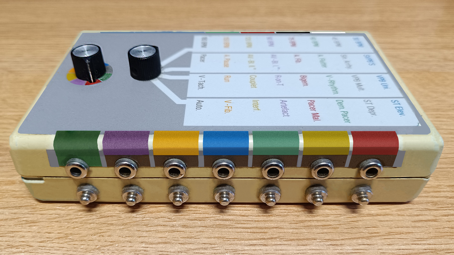 Zdjęcie przedstawia symulator elektrokardiografu, w skrócie EKG w perspektywie od prawego boku. Na obudowie znajdują się dwa pokrętła oraz cztery kolumny z oznaczeniami programów urządzenia. Na bocznej ścianie znajdują się oznaczone różnymi kolorami gniazda wyjściowe sygnału do podłączenia elektrod przyssawkowych EKG.