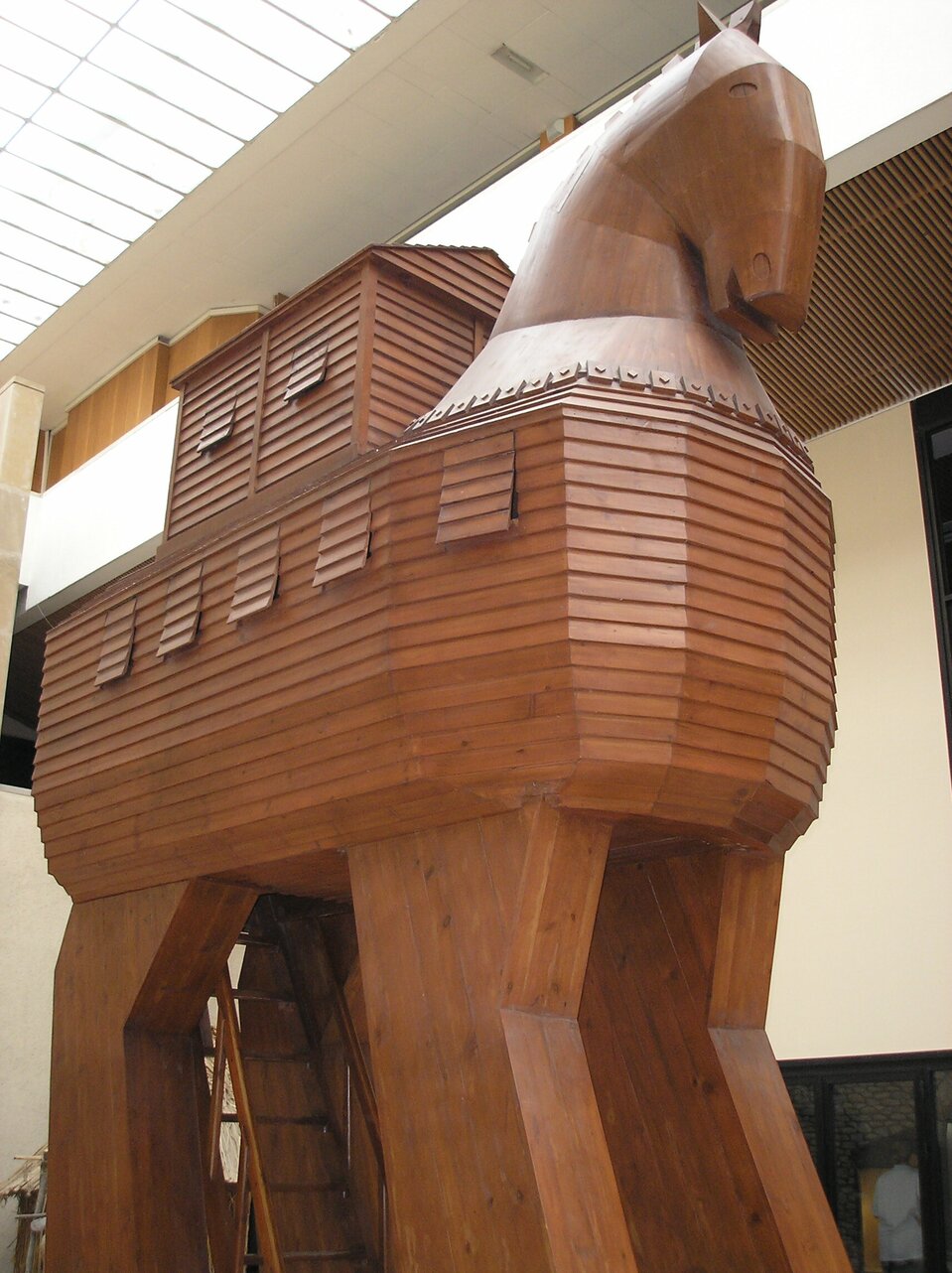 Ilustracja przedstawia monumentalnego konia trojańskiego, wykonanego z drewna. Na czterech stabilnych nogach umieszczono korpus, w którym ukryte są niewielkie okienka. Na końskim grzbiecie znajduje się drewniana nadbudówka z dwuspadowym daszkiem i dwoma okienkami. Całość dopełnia potężna końska głowa.