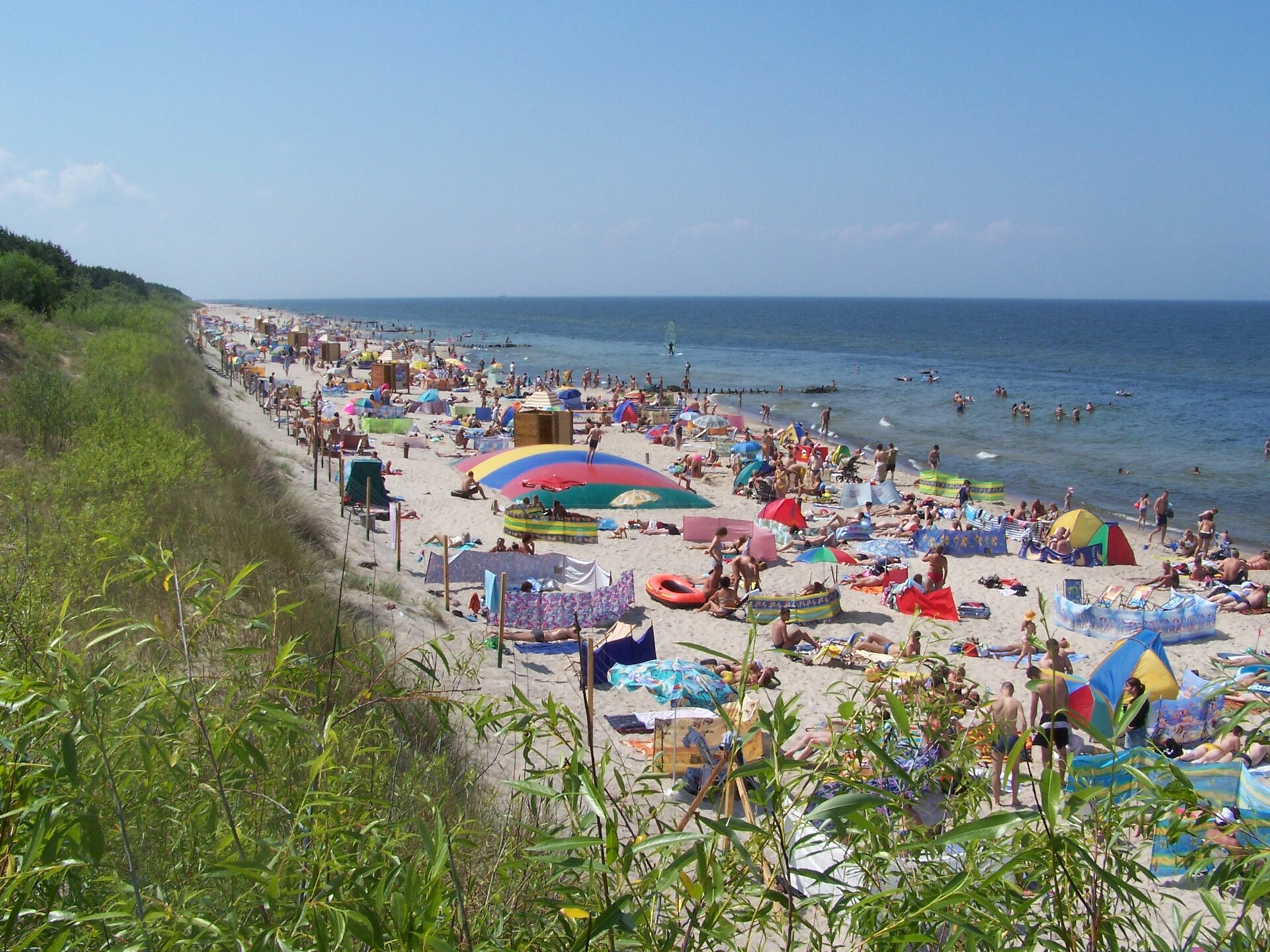 Na zdjęciu brzeg morski, piaszczysta plaża, dużo ludzi, parasole, parawany. Pas trawiastych wydm.