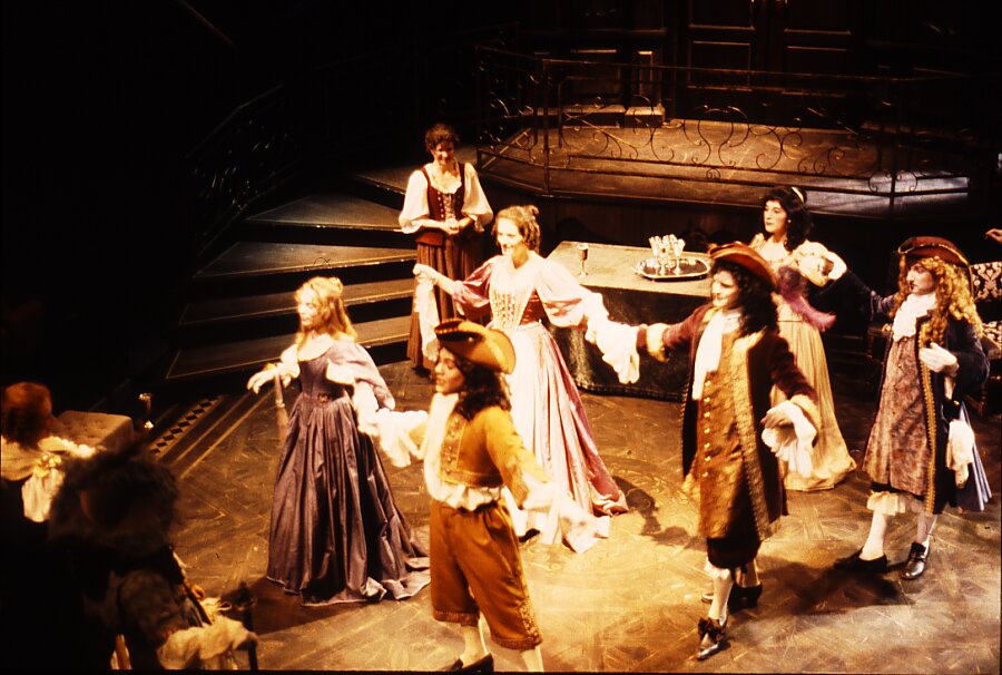 Fotografia przedstawia scenę teatralną ukazaną z perspektywy, z góry. Stoją na niej trzy pary osób tańczących menueta. Kobiety są ubrane w długie suknie. Mężczyźni mają na głowach peruki z kręconymi, długimi włosami. Na głowach mają trikorny. Ubrani są w dość długie kaftany i jasne rajtuzy.