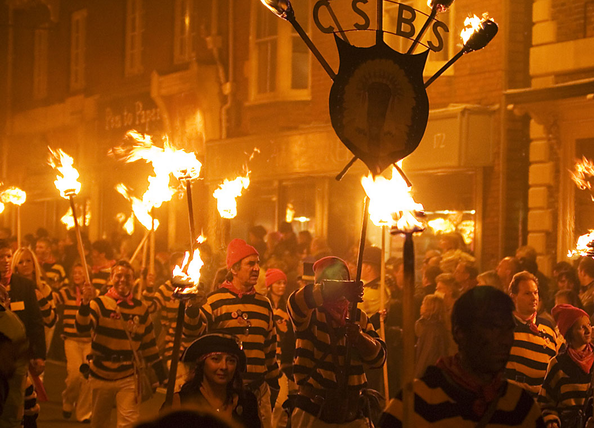 Zdjęcie przedstawia nocną paradę osób, ubranych w jednakowe stroje, czyli białe spodnie i bluzki w biało‑niebieskie paski, idących w prawo. Osoby niosą w rękach zapalone pochodnie. Na przodzie widoczna jest postać niosąca konstrukcję z tarczą i pięcioma zapalonymi pochodniami na górze, pomiędzy którymi widoczne są odwrócone w lustrzanym odbiciu litery S, B, S, C. W tle, po prawej stronie, widoczne fasady budynków oświetlone pochodniami.