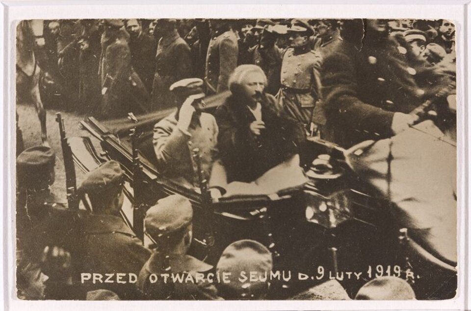 Na zdjęciu przedstawiony jest Józef Piłsudski oraz Ignacy Paderewski w drodze na otwarcie Sejmu Ustawodawczego RP w 1919 roku. Obaj politycy jadą samochodem, dokoła wita ich tłum ludzi. Zdjęcie czarno-białe.