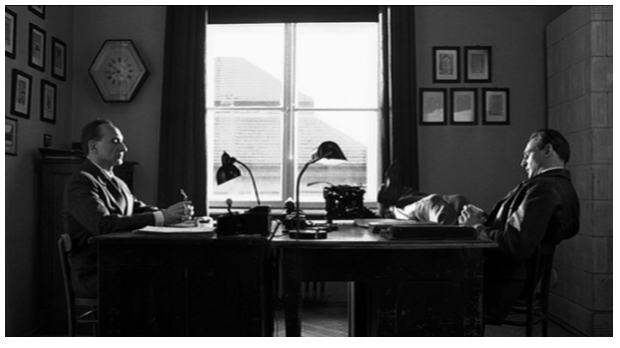 Zdjęcie przedstawia wnętrze pokoju, w którym stoją naprzeciw siebie dwa biurka. Przy biurku po prawej stronie siedzi Schindler. Jego nogi leżą na biurku. Naprzeciw niego jest Stern. Siedzi wyprostowany.