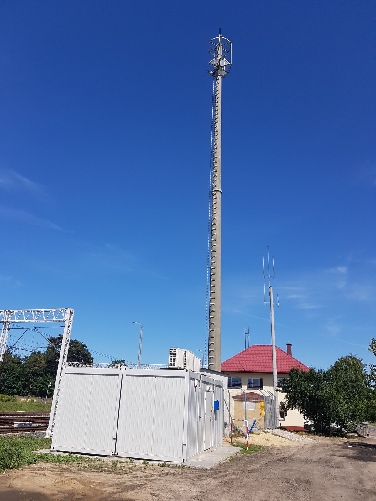 Zdjęcie przedstawia maszt radiowy GSM‑R. Widoczny jest wysoki masz stojący obok torów kolejowych. Na szczycie masztu znajduje się platforma z antenami. U stóp masztu stoi mały budynek z blachy.