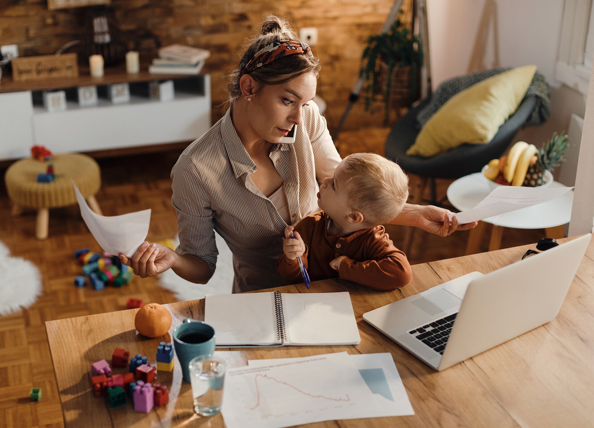 Zdjęcie przedstawia młodą kobietę siedzącą za biurkiem, na którym stoi otwarty laptop. Kobieta ma ciemne włosy związane na czubku głowy, opaskę na włosach i jest ubrana w koszulę w paski. Na kolanach kobiety siedzi małe dziecko. Dziecko ma jasne krótkie włosy i jest ubrane w brązowy sweter. Kobieta przy lewym uchy ma telefon, który przytrzymuje lewym barkiem. Jej głowa jest lekko przechylona w prawo. Kobieta spogląda na dziecko. Obie ręce ma rozłożone na boki i trzyma w nich kartki. Dziecko ma głowę lekko uniesioną i patrzy na kobietę. W prawej dłoni trzyma długopis. Na biurku, na lewo od laptopa widoczny jest otwarty notes, jakieś wydruki, kubek, szklanka, kolorowe klocki i mandarynka. W tle widoczne jest wyposażenie pokoju: mały biały stolik, na którym stoją owoce w misce, fotel z poduszkami, niski taboret, szafla, a na podłodze klocki. 