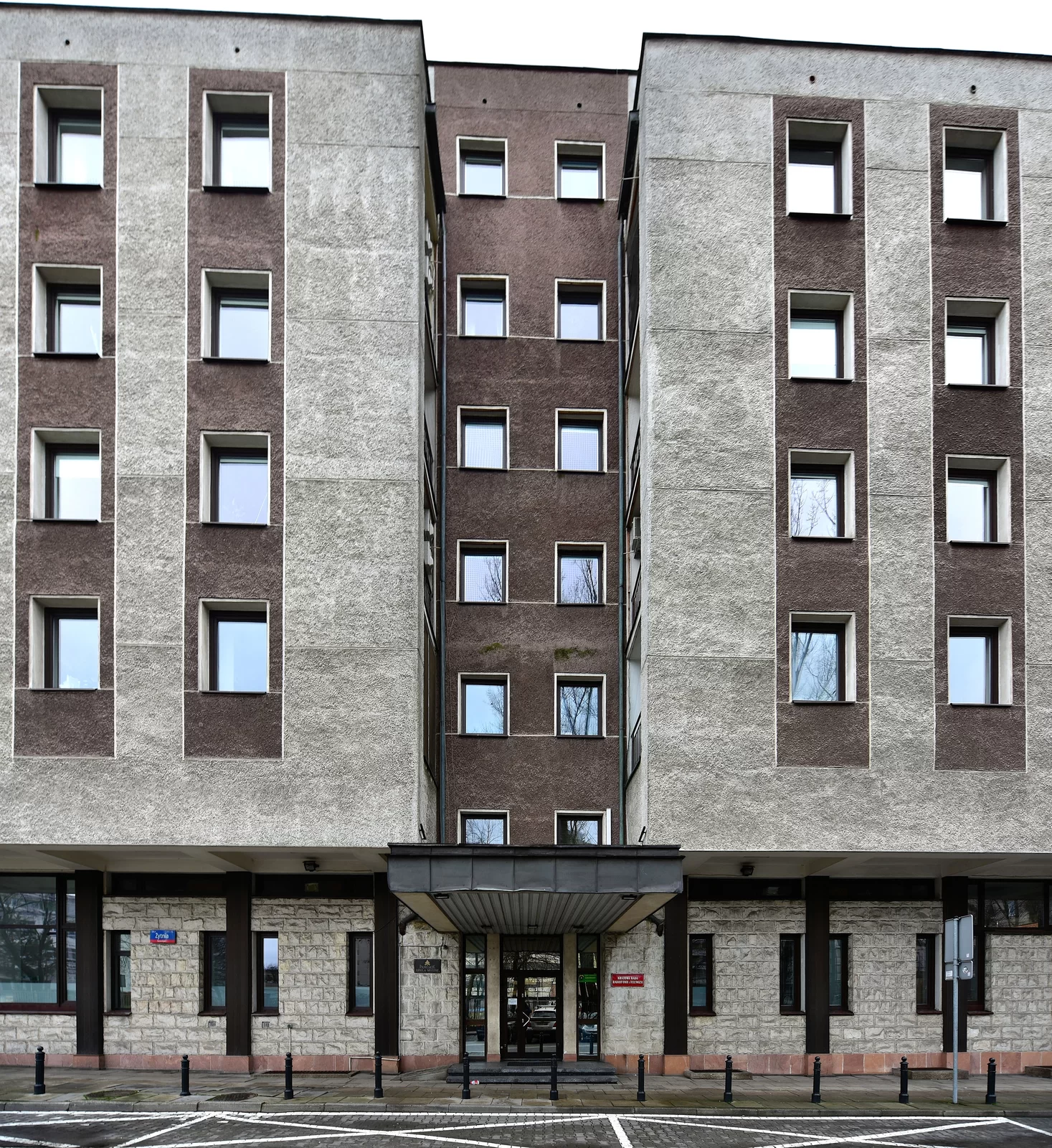 Zdjęcie przedstawia duży, czteropiętrowy budynek, w którym mieści się Siedziba Krajowej Rady Radiofonii i Telewizji w Warszawie. Do budynku prowadzi duże wejście osłonięte daszkiem. Elewacja jest szara, przy oknach jest ona ciemnobrązowa.