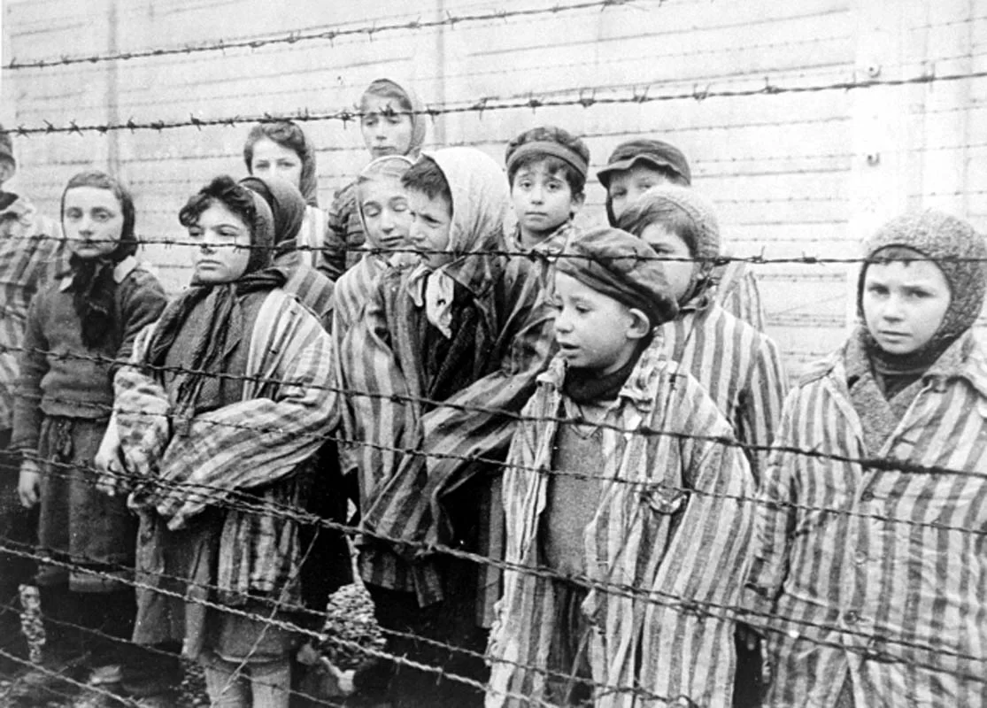 Czarno‑białe zdjęcie przedstawia więźniów obozu. Chłopcy mają na głowie czapki. Są ubrani w koszule, swetry i spodnie. Natomiast dziewczęta mają chustki na głowie. Są ubrane w sukienki do kolan i rajstopy. Wszystkie dzieci są ubrane w pasiaste koszule z długim rękawem. Od strony widza dzieci stoją za drutem kolczastym.