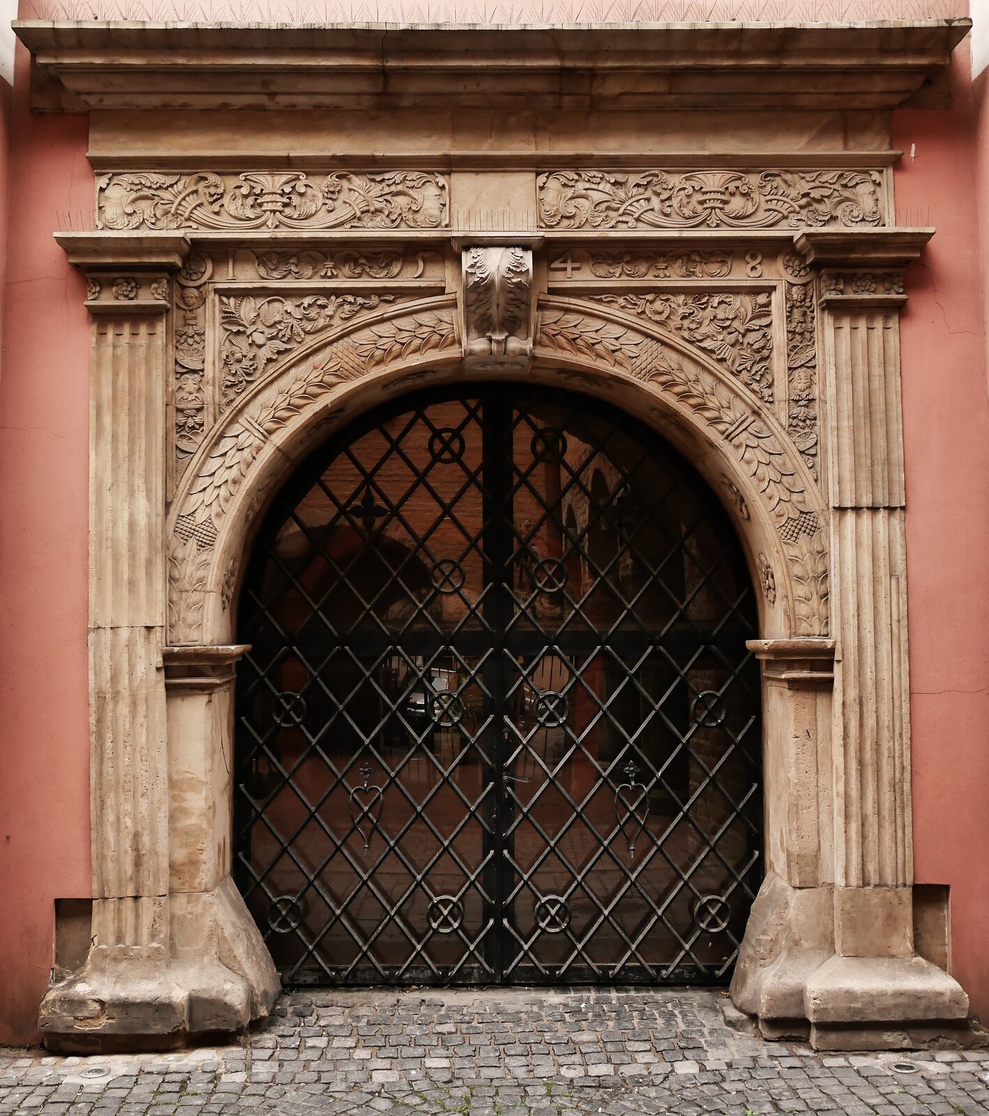 Zdjęcie przedstawia nieduży portal o łagodnym łuku. Jest wykonany z jasnego kamienia. Zdobiony roślinnymi motywami. W portalu wstawiono czarną, ażurową bramę.