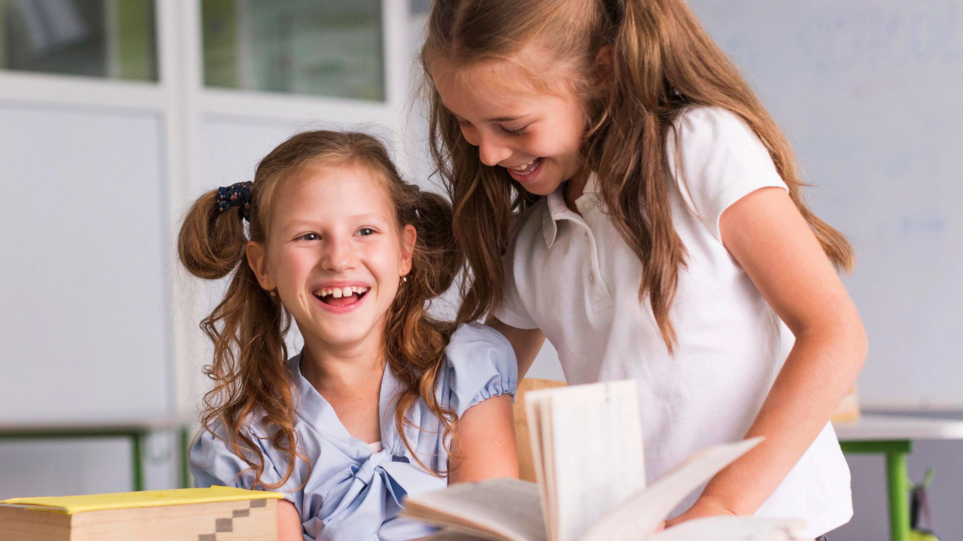 Zdjęcie przedstawia dwie uśmiechnięte nastoletnie dziewczyny z długimi włosami. Dziewczyna po lewej siedzi, a druga stoi. Przed dziewczynami na stoliku znajduje się otwarta książka.