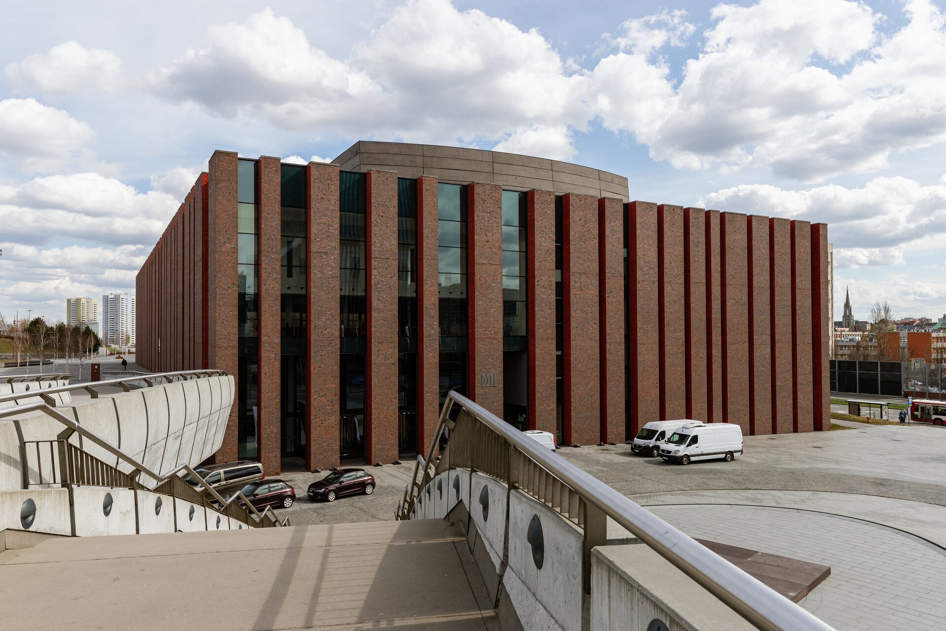 Fotografia przedstawia nowoczesny budynek o brązowej elewacji. Na pierwszym planie znajdują się schody prowadzące na betonowy plac, na którym znajduje się fontanna oraz dwa białe samochody dostawcze, dwa bordowe samochody osobowe oraz jedna brązowa furgonetka. Na drugim planie znajduje się budynek z brązowej cegły, widoczne są dwie ściany poprzecinane pionowymi szczelinami, w dwóch największych widoczne są okna. W tle znajdują się wieżowce. 