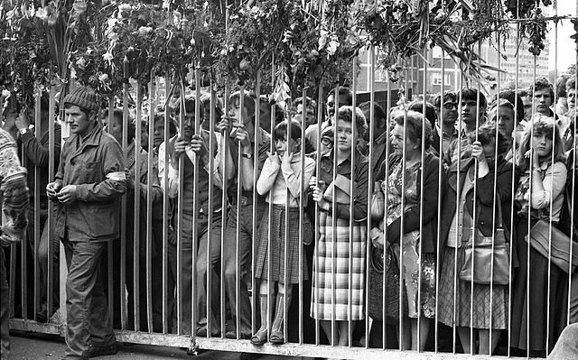 Zdjęcie przedstawia tłum ludzi stojących za metalowym ogrodzeniem Stoczni Gdańskiej. Są tu przede wszystkim kobiety.